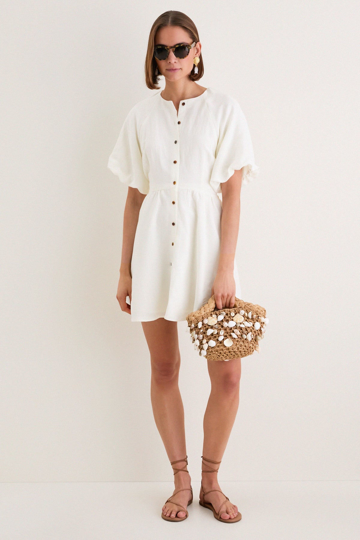 Woman wearing the classic Star White Bastille Short Dress, with effortless puffed sleeves and a button-front, accessorized with a woven straw tote, embodying high-end, fun style.