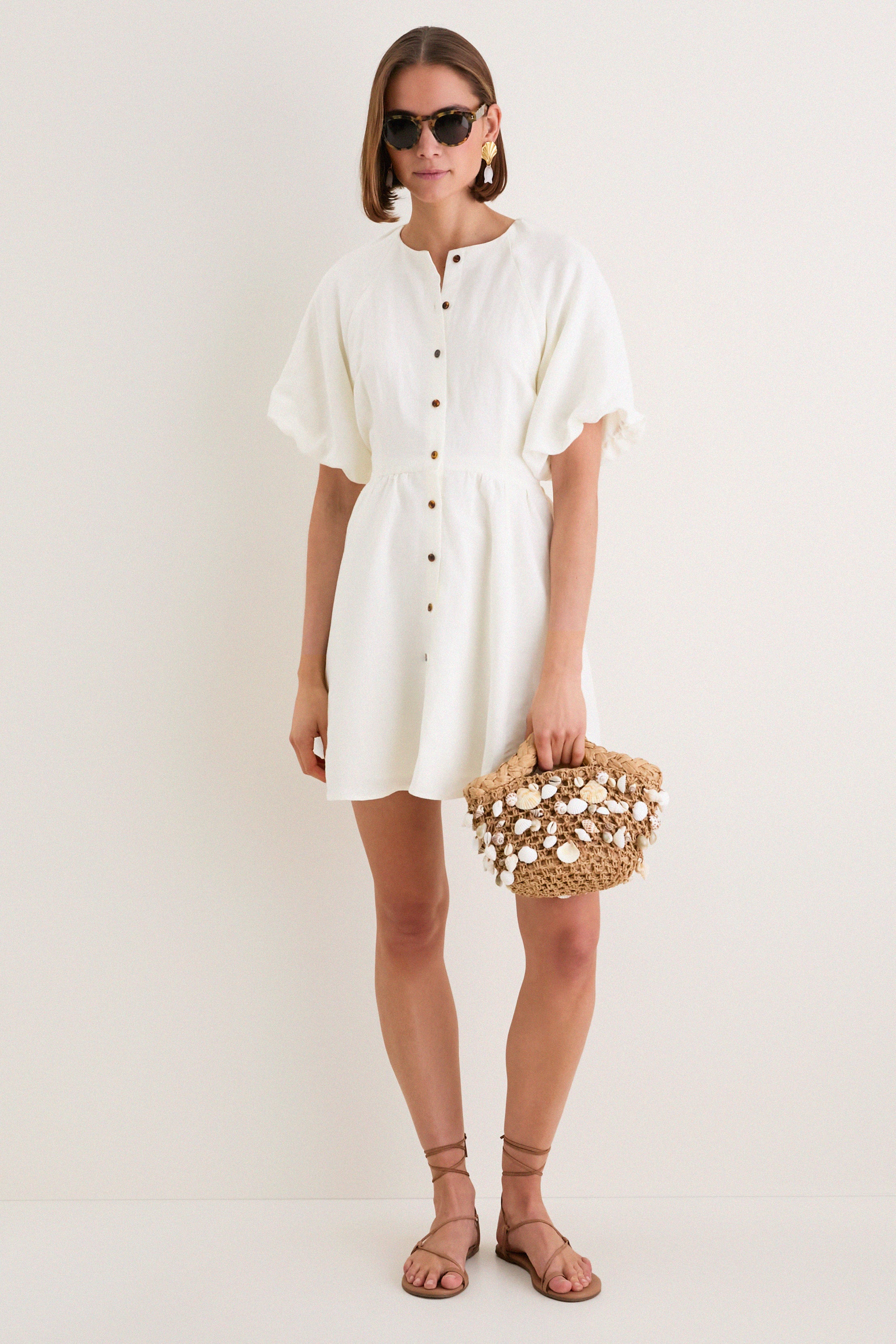 Woman wearing the classic Star White Bastille Short Dress, with effortless puffed sleeves and a button-front, accessorized with a woven straw tote, embodying high-end, fun style.