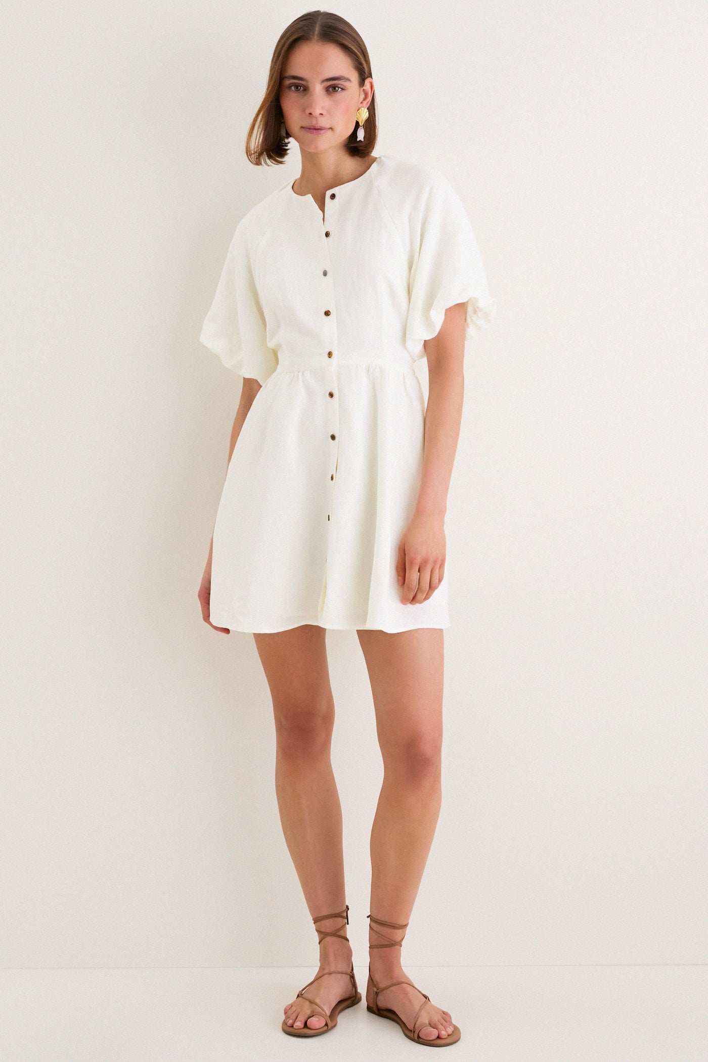 Model in classic, white Star White Bastille Short Dress with flutter sleeves, high neckline, self-tie waist, paired with tan sandals and statement earrings. Effortless, high-end style.
