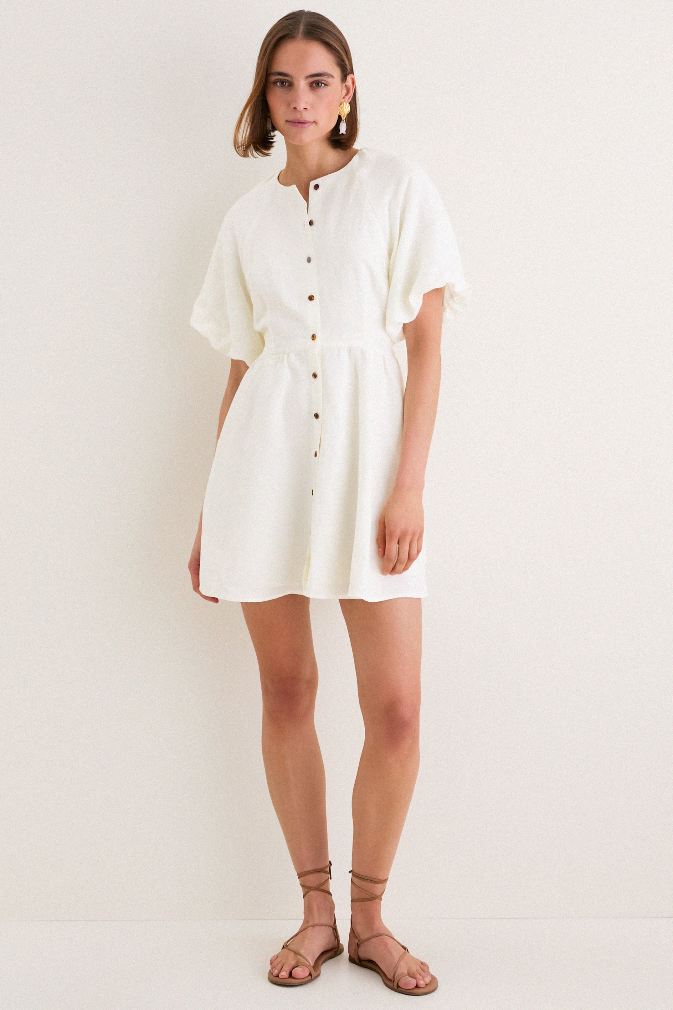 Model in classic, white Star White Bastille Short Dress with flutter sleeves, high neckline, self-tie waist, paired with tan sandals and statement earrings. Effortless, high-end style.