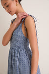 Young woman adjusts tie strap on a classic Navy Gingham Sloan Maxi Dress, showcasing its effortless, high-end design with a fun and romantic touch.