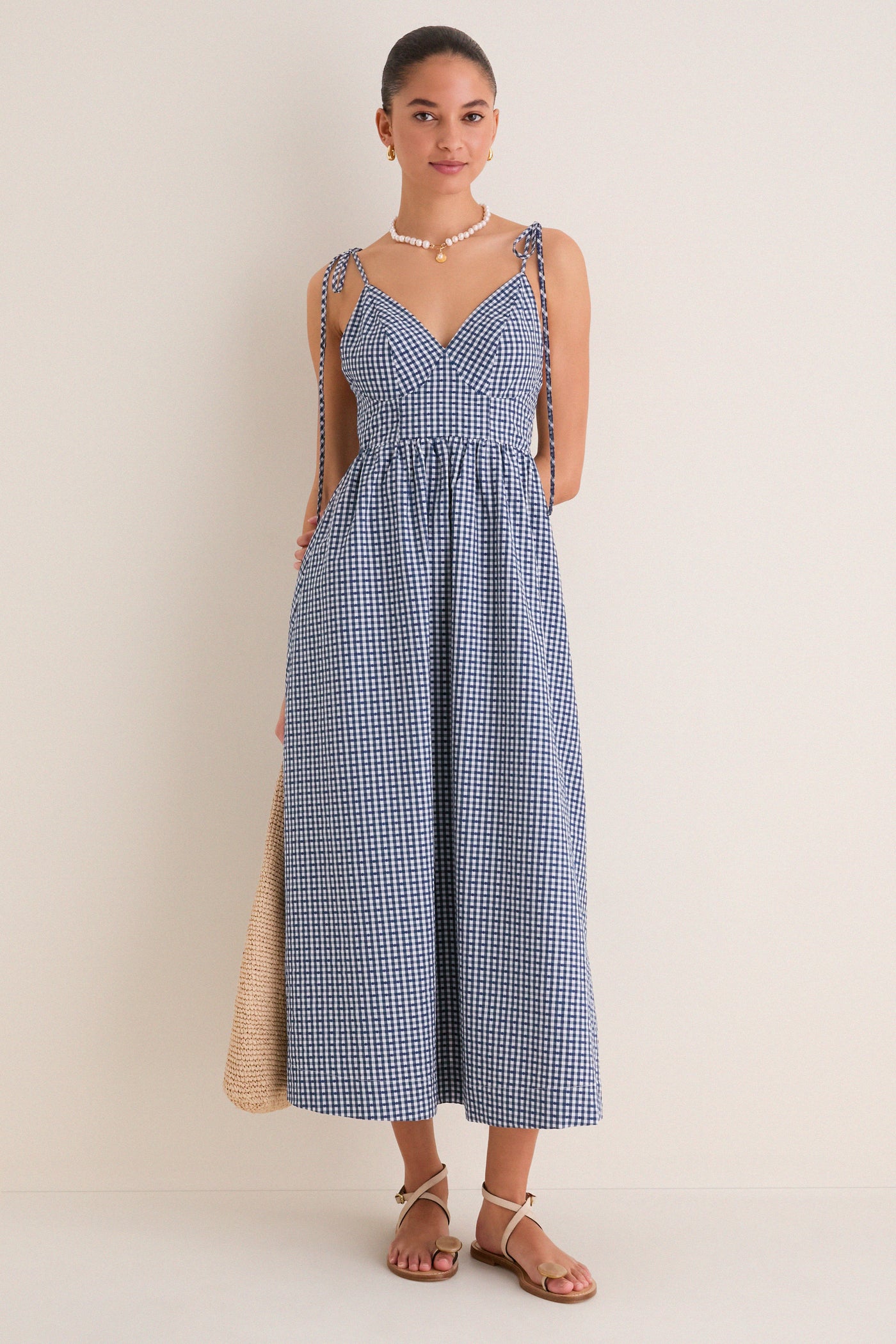 Young woman in a classic, effortless Navy Gingham Sloan Maxi Dress with tie straps, accessorized with pearls and sandals, holding a straw tote, embodying high-end, fun style.
