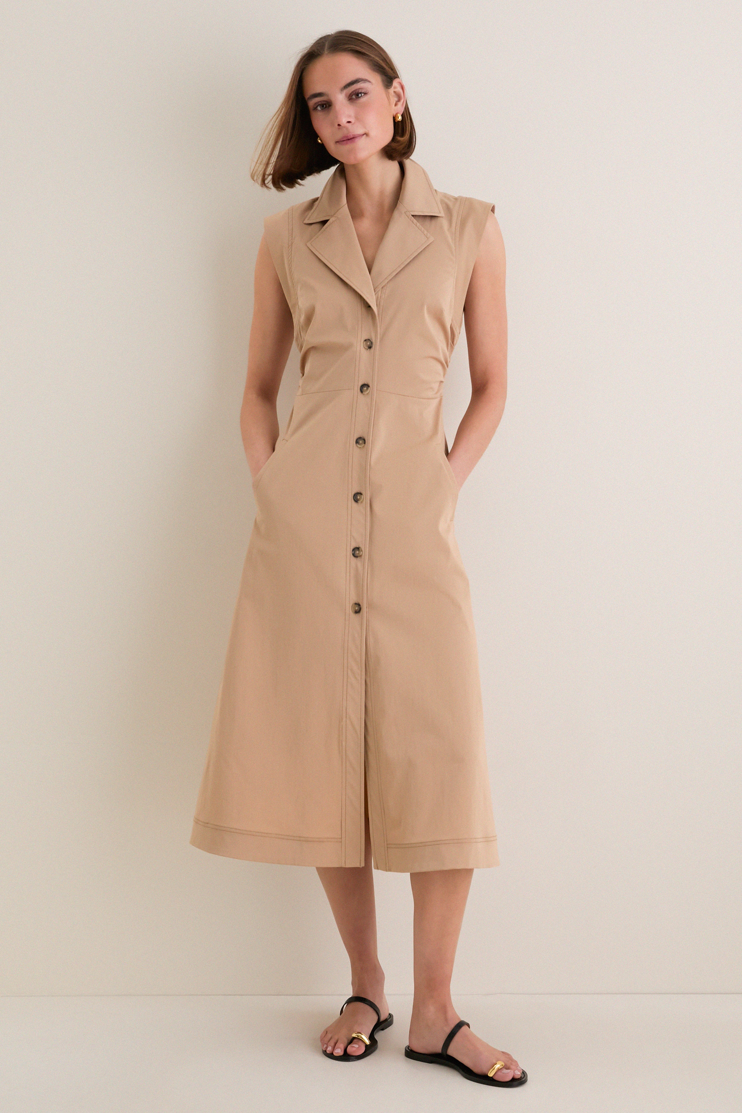 Young woman models the classic Khaki Orla Dress, a sleeveless, button-down midi with side pockets, showcasing its effortless, high-end style paired with fun black sandals.