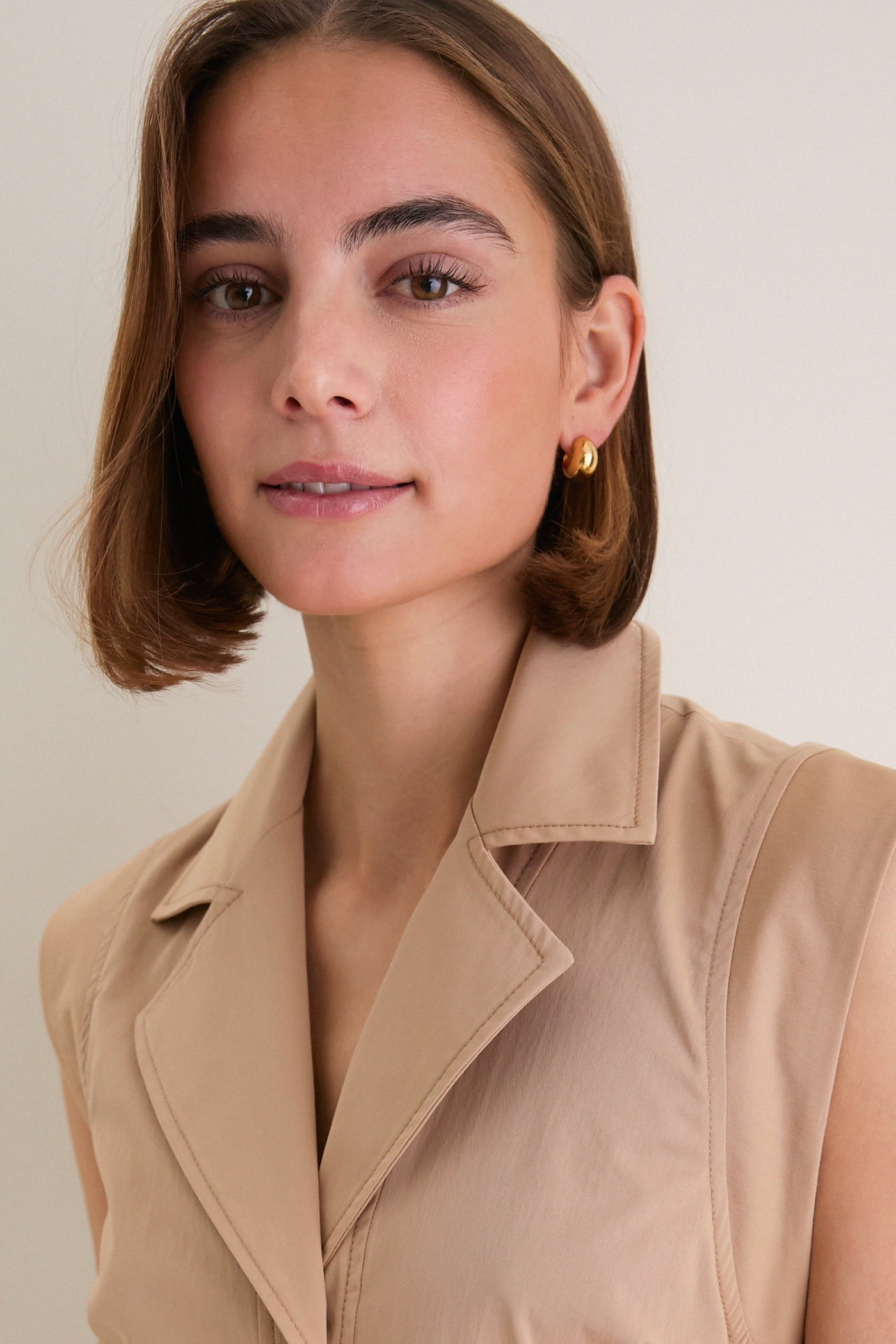 Young woman wearing the classic Khaki Orla Dress with gold hoop earrings, showcasing its effortless fit-and-flare design, cap sleeves, and tailored detailing. High-end, fun, and versatile style.