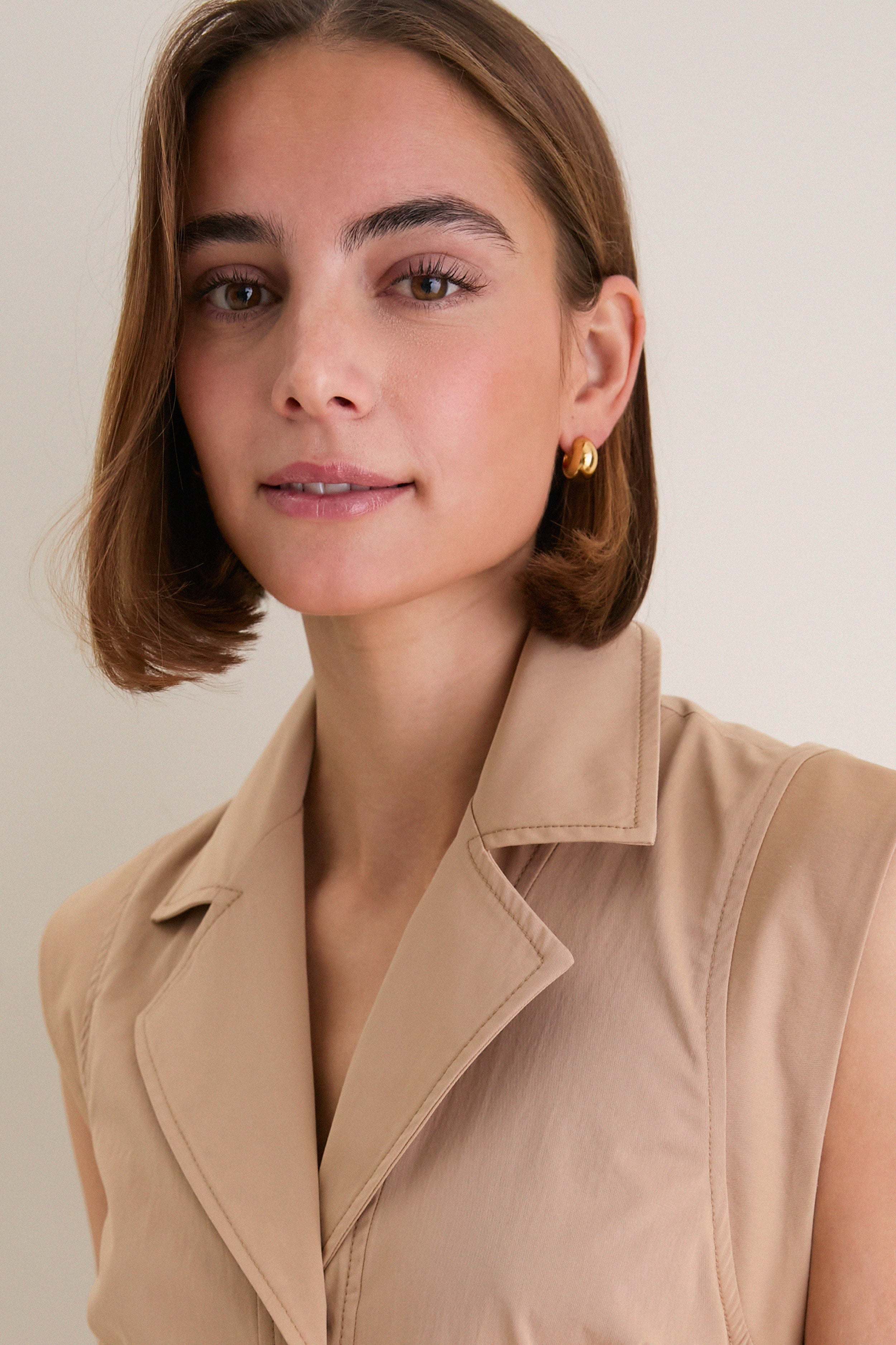 Young woman wearing the classic Khaki Orla Dress with gold hoop earrings, showcasing its effortless fit-and-flare design, cap sleeves, and tailored detailing. High-end, fun, and versatile style.