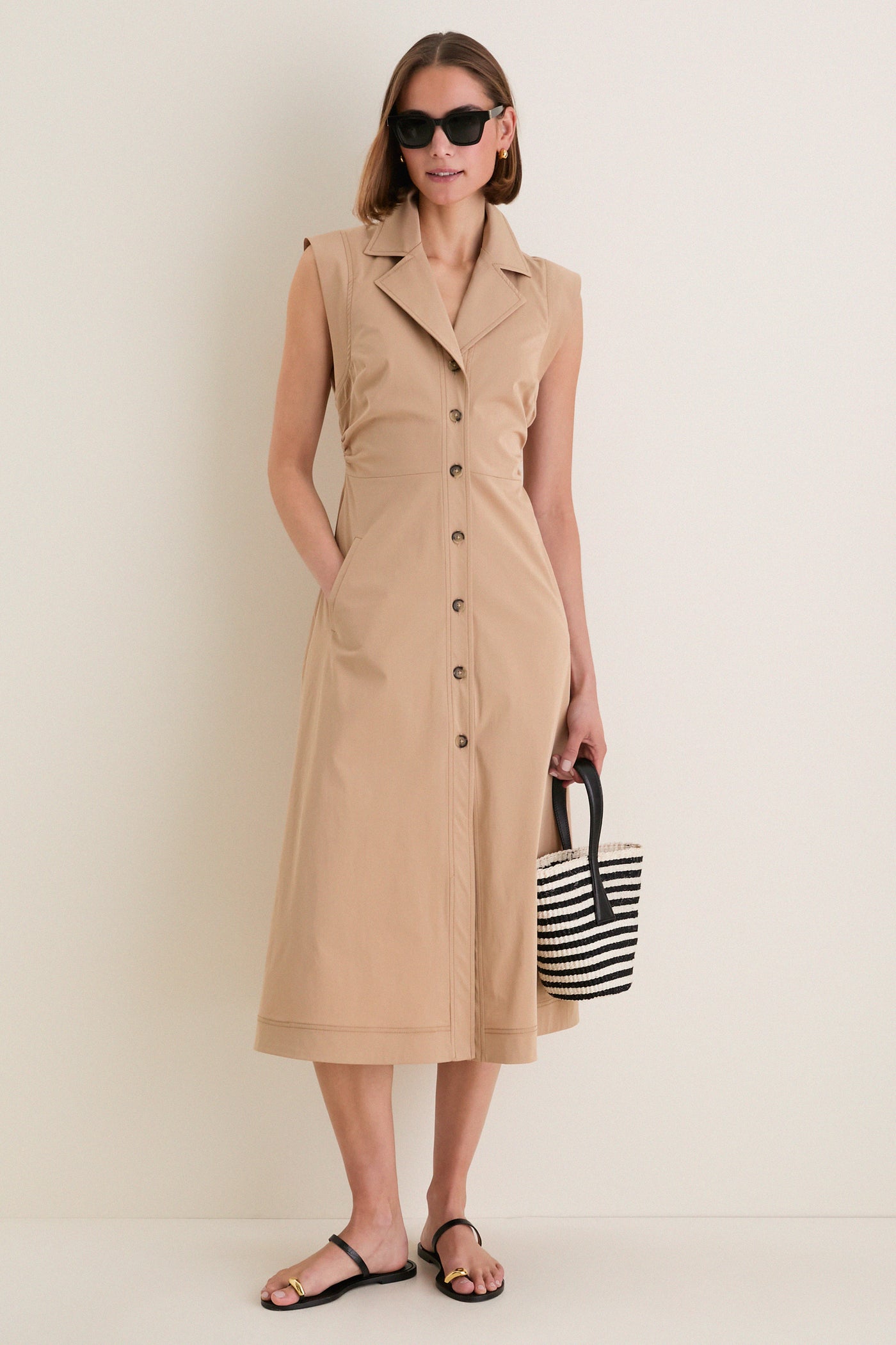Woman wearing the classic Khaki Orla Dress with side ruching and cap sleeves, styled effortlessly with black sandals, oversized sunglasses, and a striped tote bag.