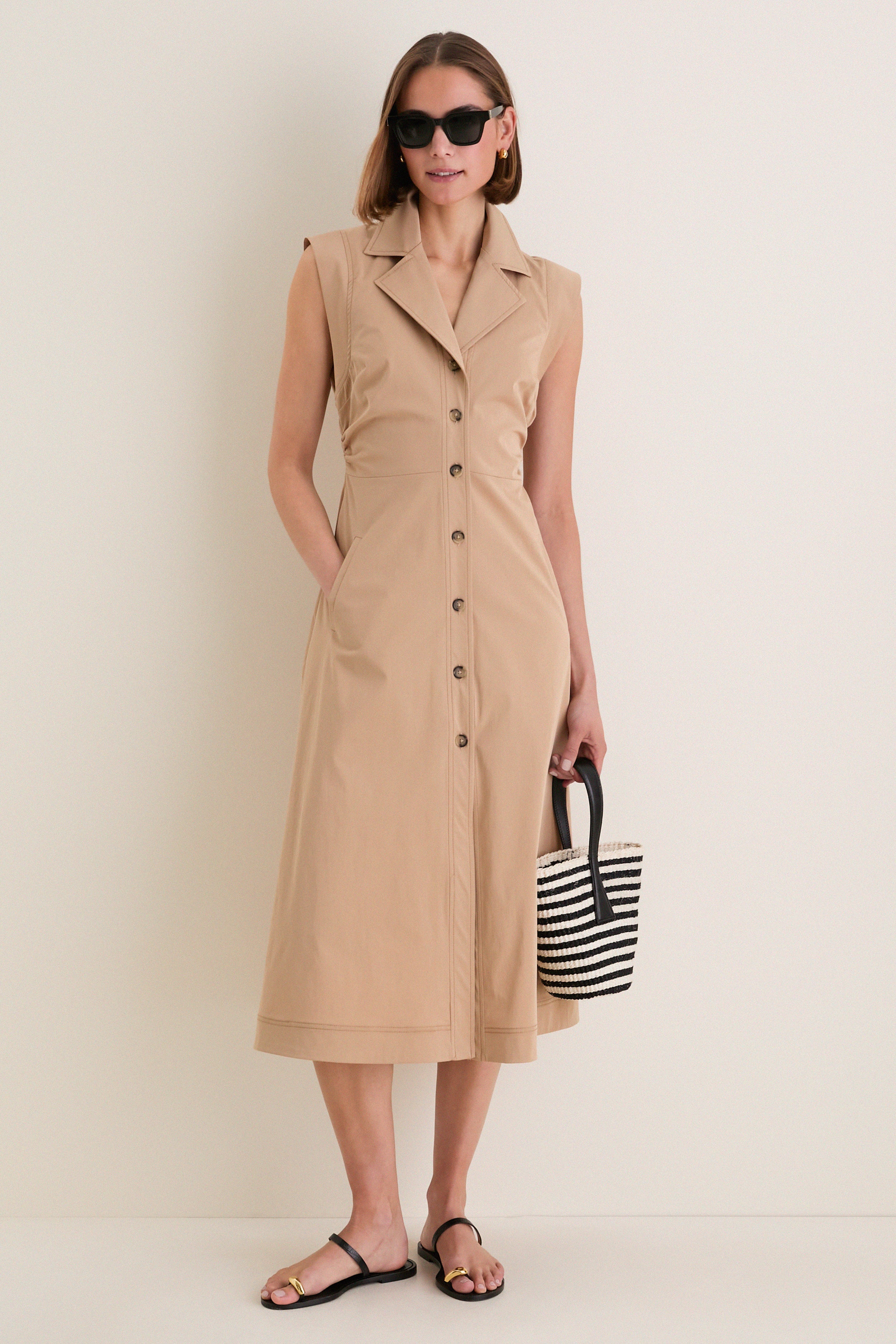 Woman wearing the classic Khaki Orla Dress with side ruching and cap sleeves, styled effortlessly with black sandals, oversized sunglasses, and a striped tote bag.