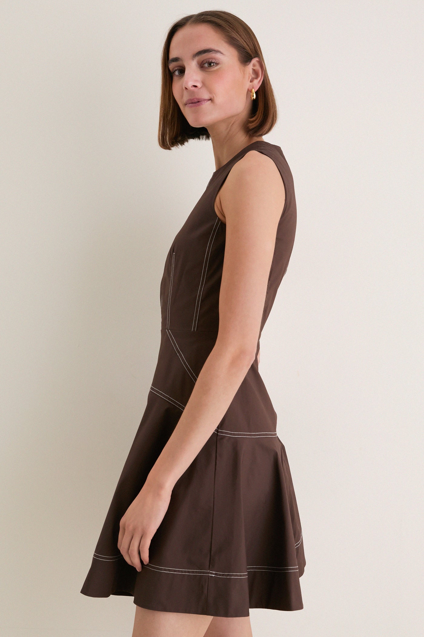Young woman models the Dark Mocha Mini Karrigan Dress, showcasing its classic, sleeveless fit-and-flare design with effortless contrast topstitching and high-end, playful style.