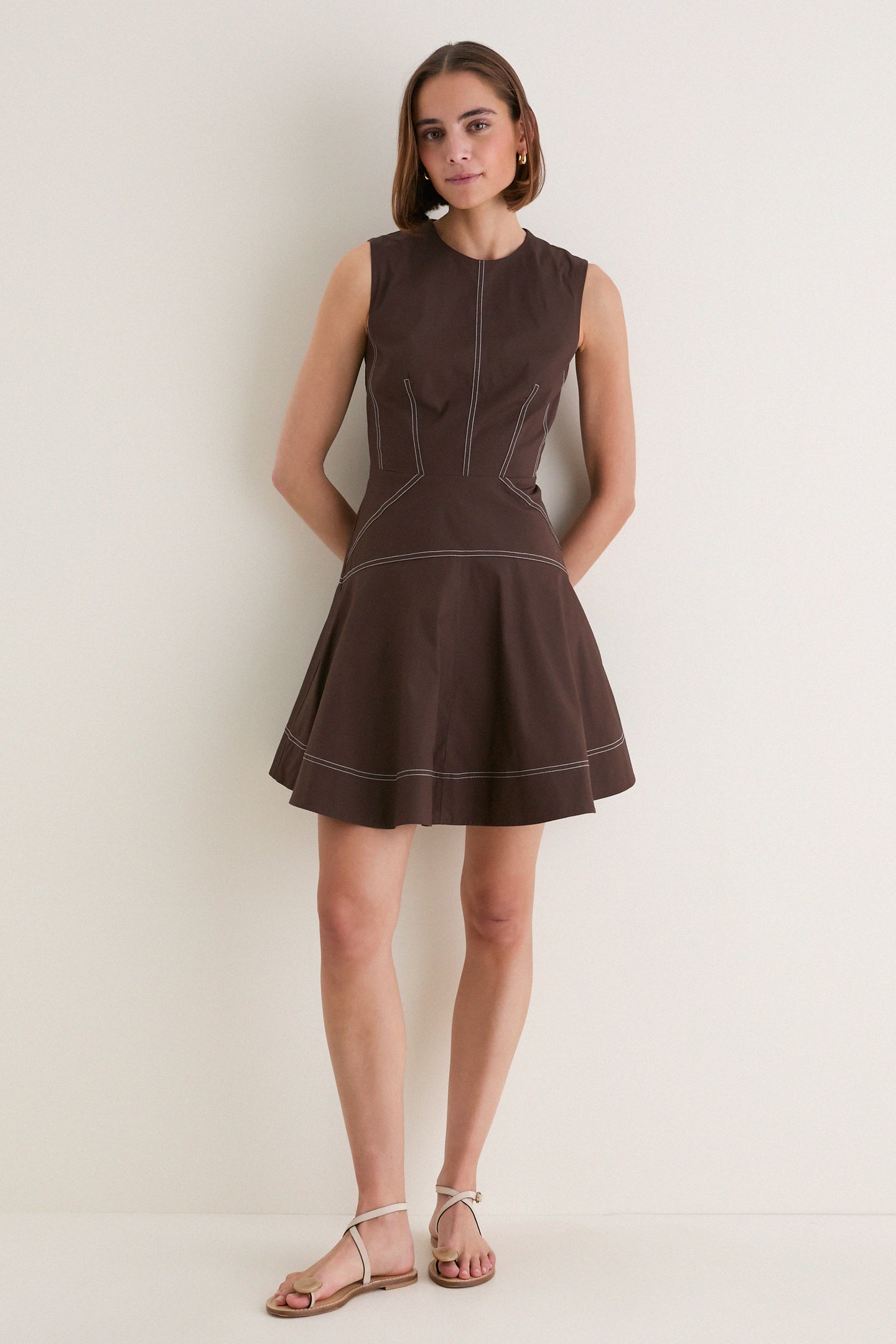 Young woman in a Dark Mocha Mini Karrigan Dress, a sleeveless, fit-and-flare style with classic crew neckline and playful contrast topstitching, embodying effortless high-end fashion.
