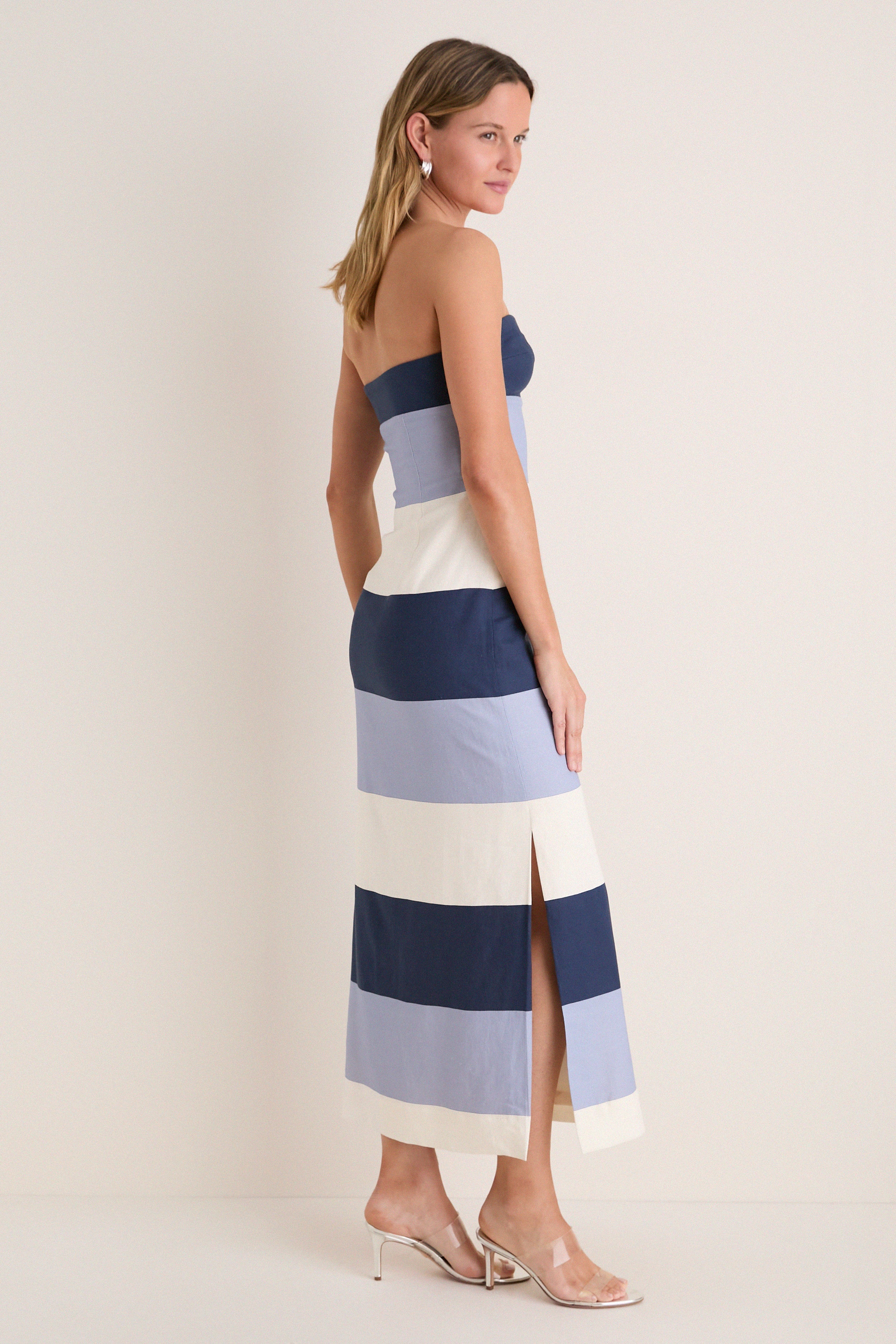 Woman in a classic Blue Stripes Sono Maxi Dress with high-end nautical design, effortless silhouette, thigh-high slit, and clear-strap heels, exuding fun sophistication.