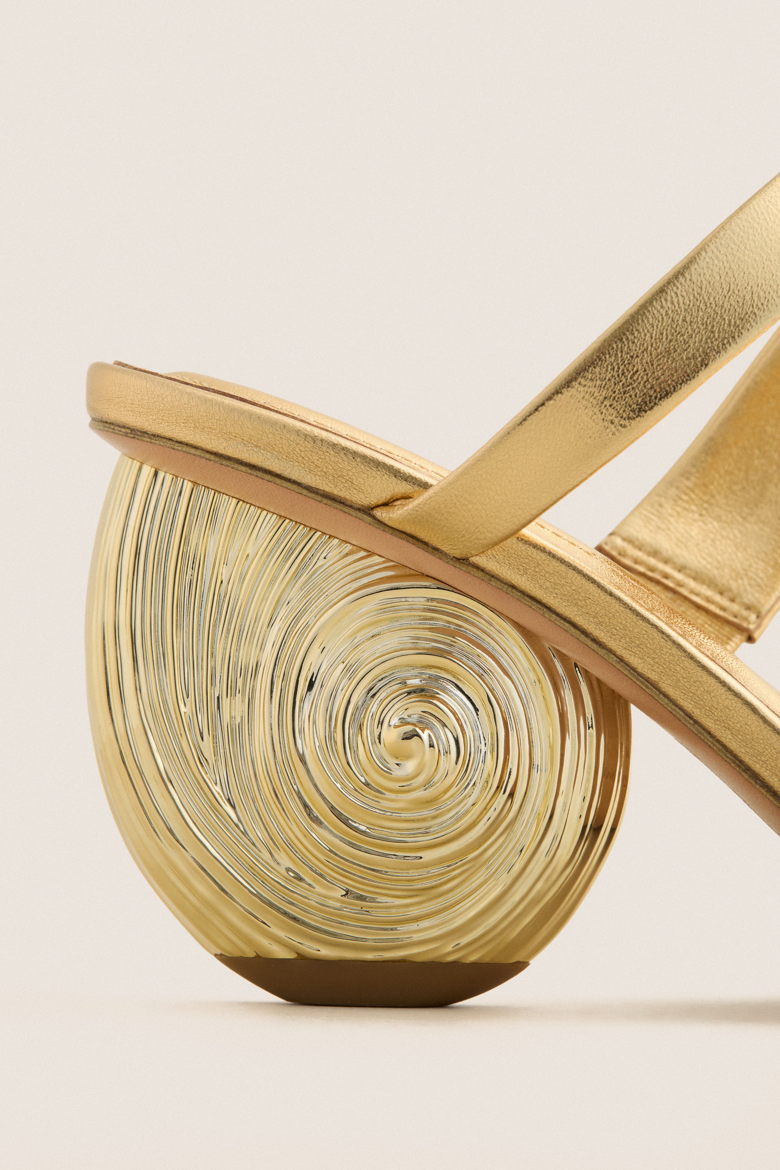 Gold Marion Metallic Shell Heels with a classic, sculptural half-circle spiral heel and an effortless upper strap, embodying high-end elegance and fun sophistication.