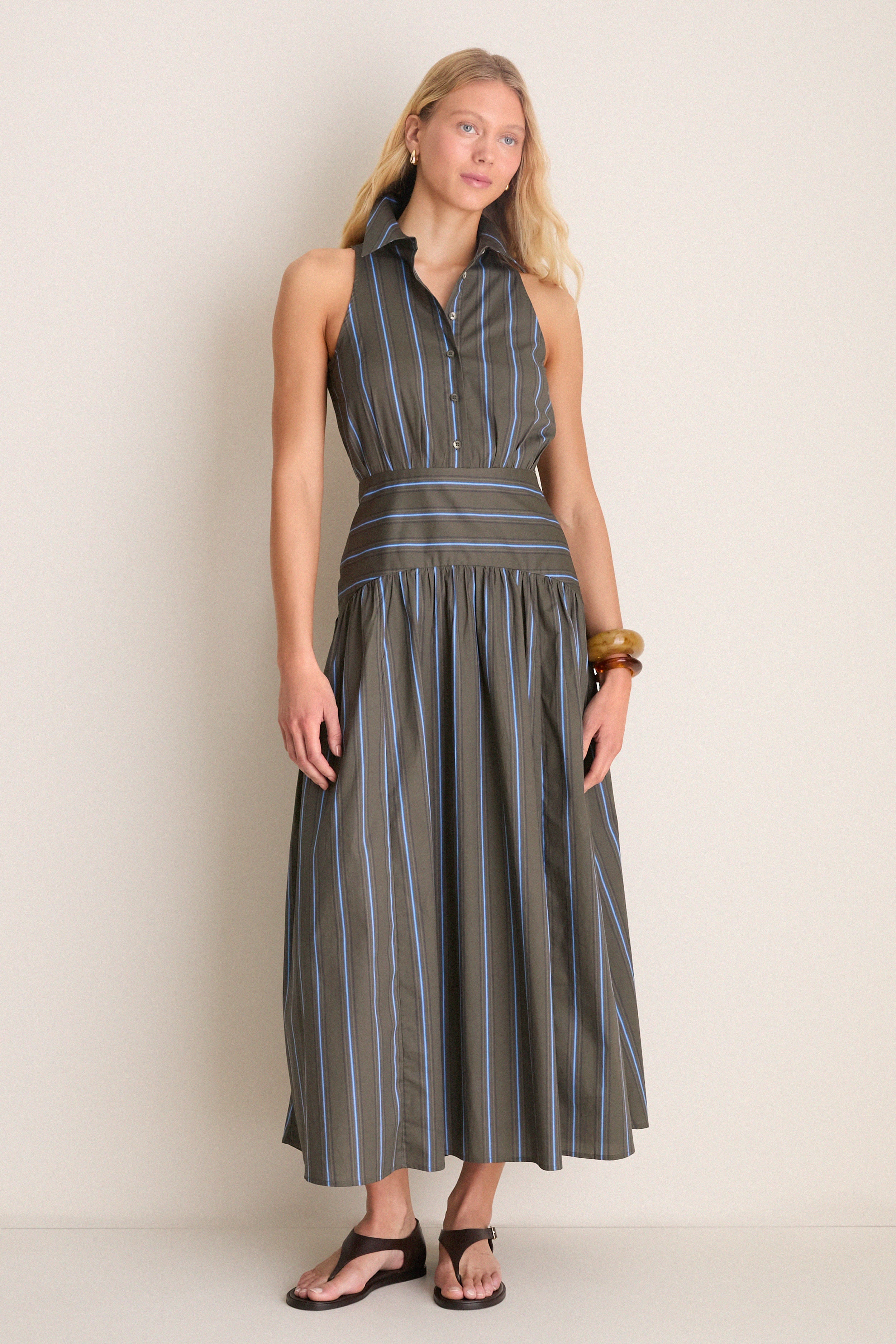 Young woman in Huntington Stripe Romy Dress: a sleeveless, classic maxi with button front and fitted waistband, styled effortlessly with brown sandals and chunky bracelets.