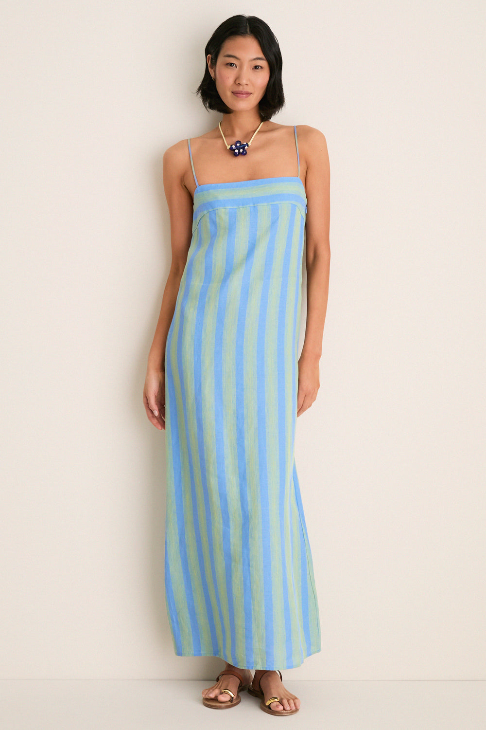 Woman models Seaport Stripe Laura Linen Dress, a classic, high-end maxi with blue and green stripes, spaghetti straps, and a square neckline. She complements it with gold-accent sandals.
