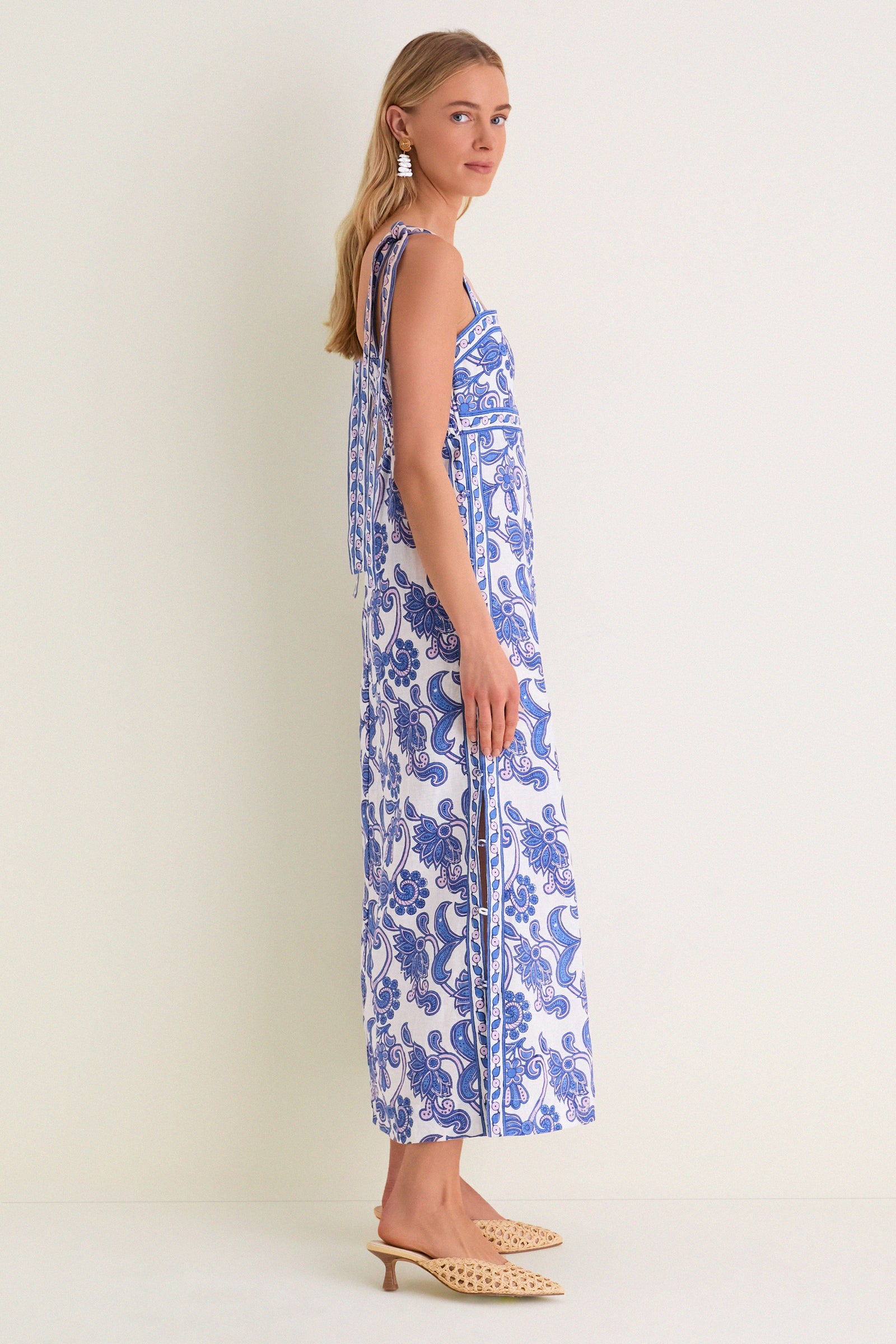 Young woman in Brocade Azul Tali Maxi Dress, a classic white dress with blue paisley-floral print, showcasing effortless style with fun tie-shoulder straps and high-end woven mules.