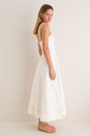 Model in profile wearing the Off White Fiona Dress, a classic, ankle-length piece with spaghetti straps and a flared skirt, paired with effortless gold sandals.