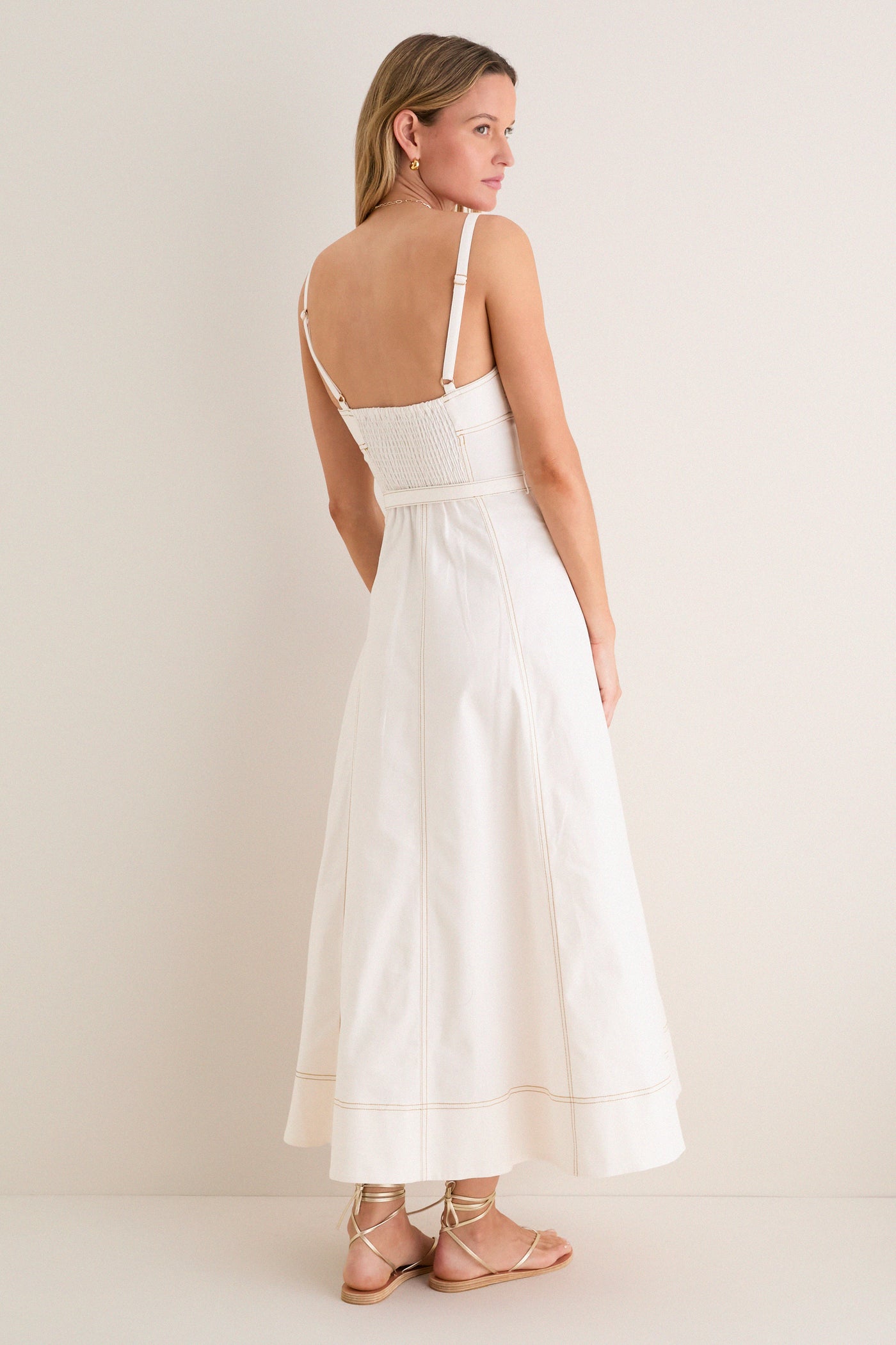 Woman models the classic Off White Fiona Dress, featuring an effortless fitted smocked bodice, A-line skirt, adjustable straps; paired with fun, high-end gold-tone sandals and hoop earrings.