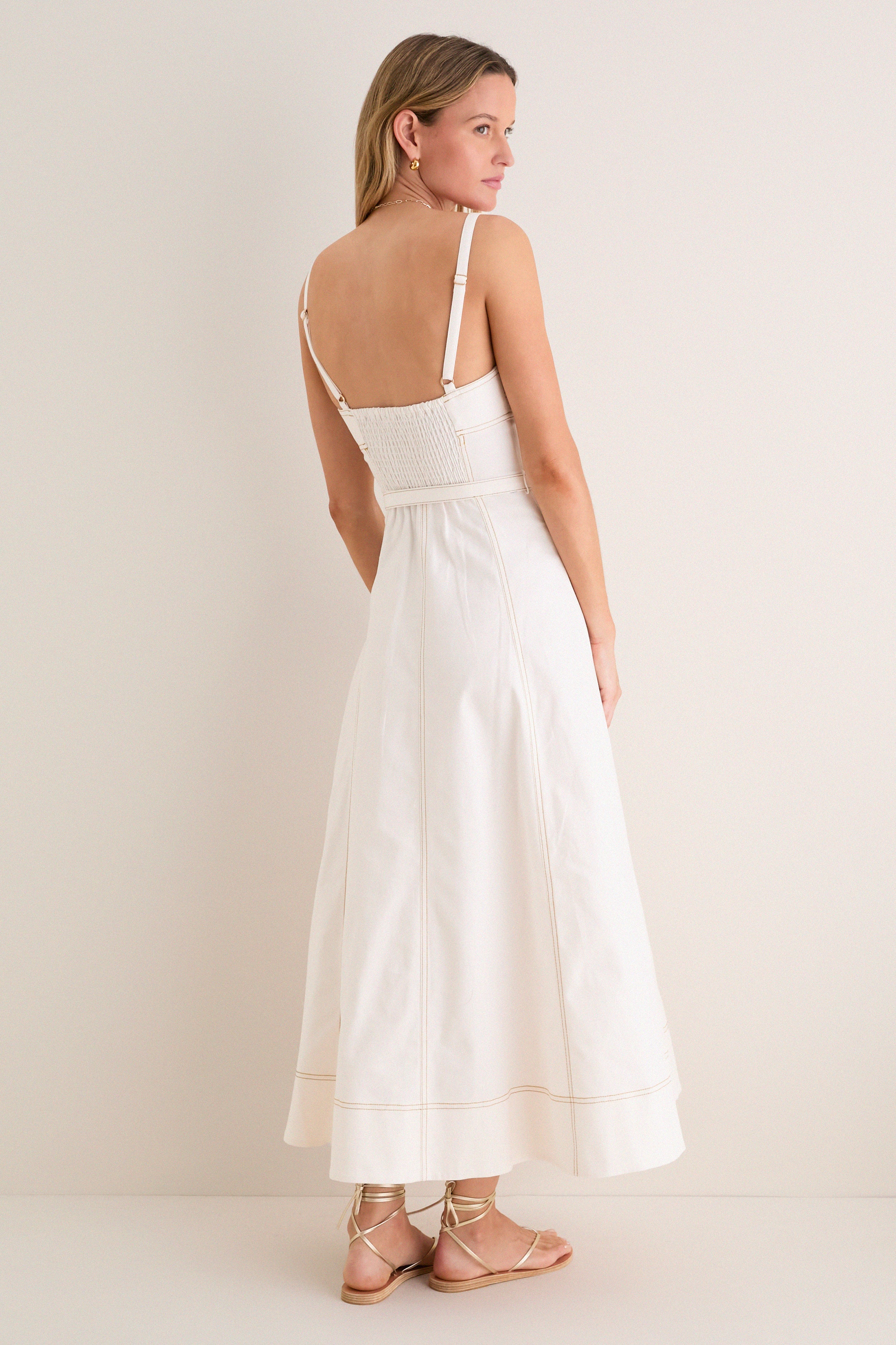 Woman models the classic Off White Fiona Dress, featuring an effortless fitted smocked bodice, A-line skirt, adjustable straps; paired with fun, high-end gold-tone sandals and hoop earrings.