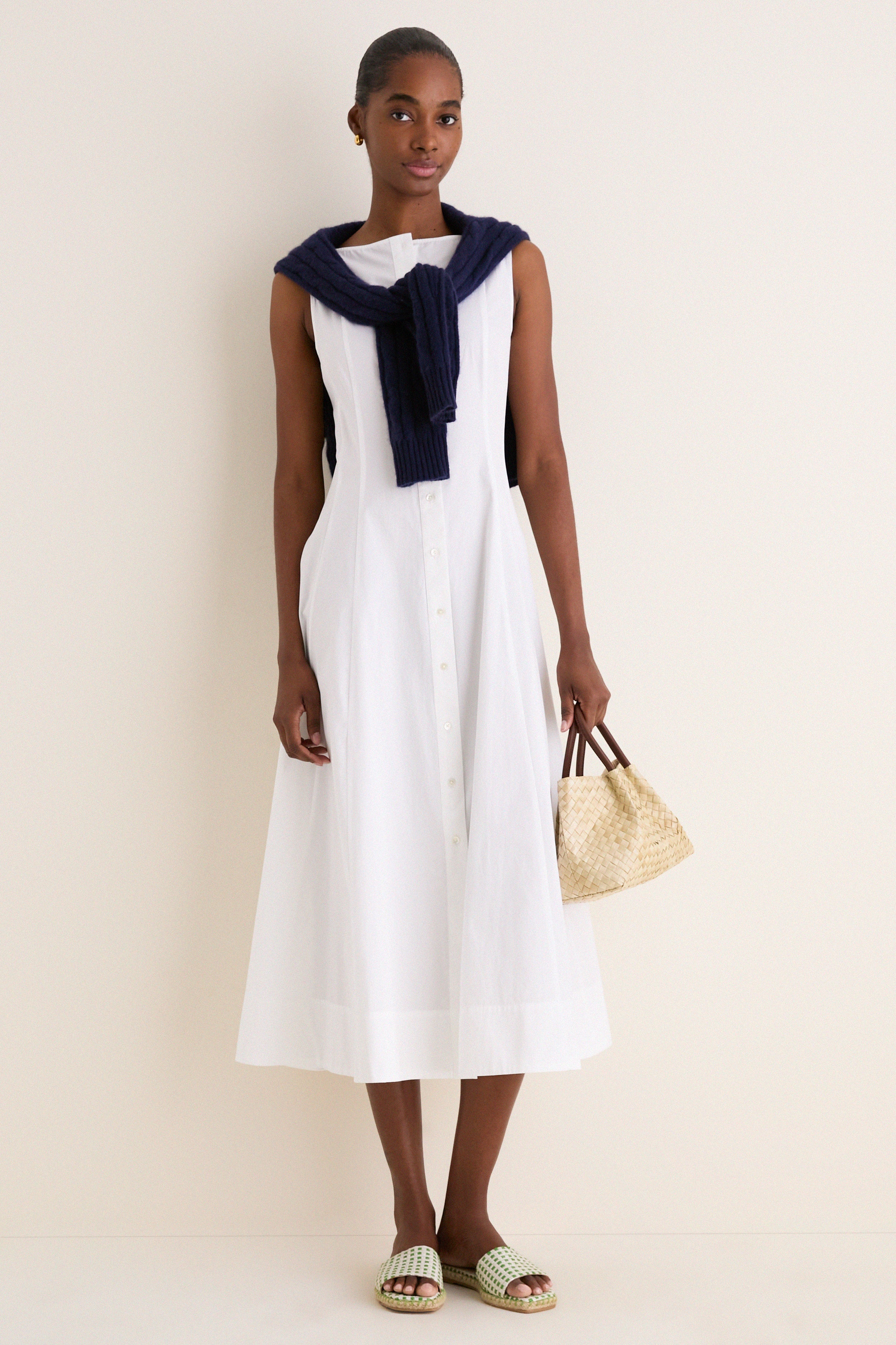 Young woman in a classic, sleeveless White Cotton Boat Neck Dress, draped with a navy sweater, carrying a straw tote, embodying an effortless, high-end resort style.