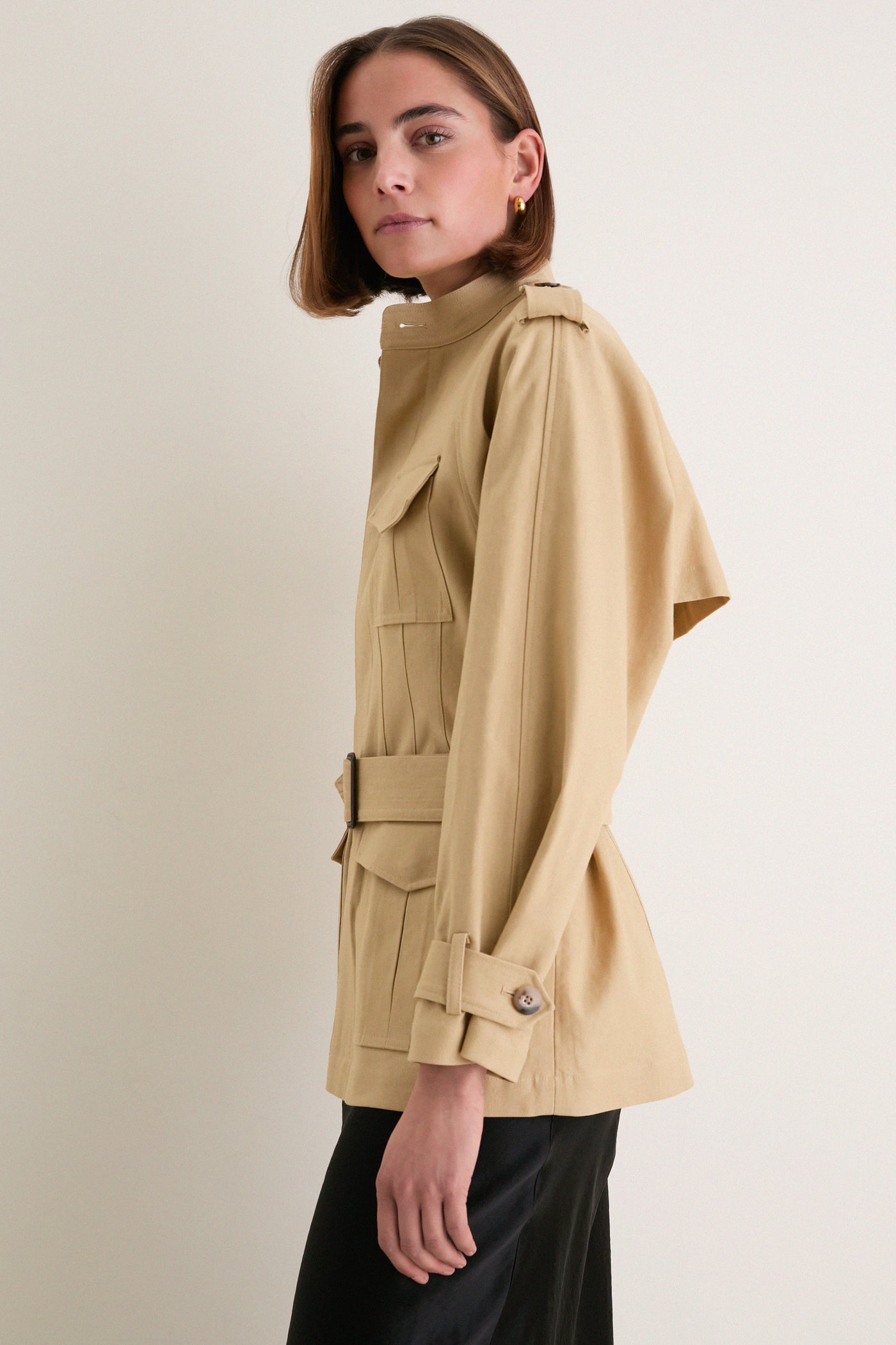 Young woman in profile modeling the Classic Camel Cotton Field Jacket, featuring a spread collar, buttoned placket, and cinched waist, paired with black trousers. Effortless and high-end style.
