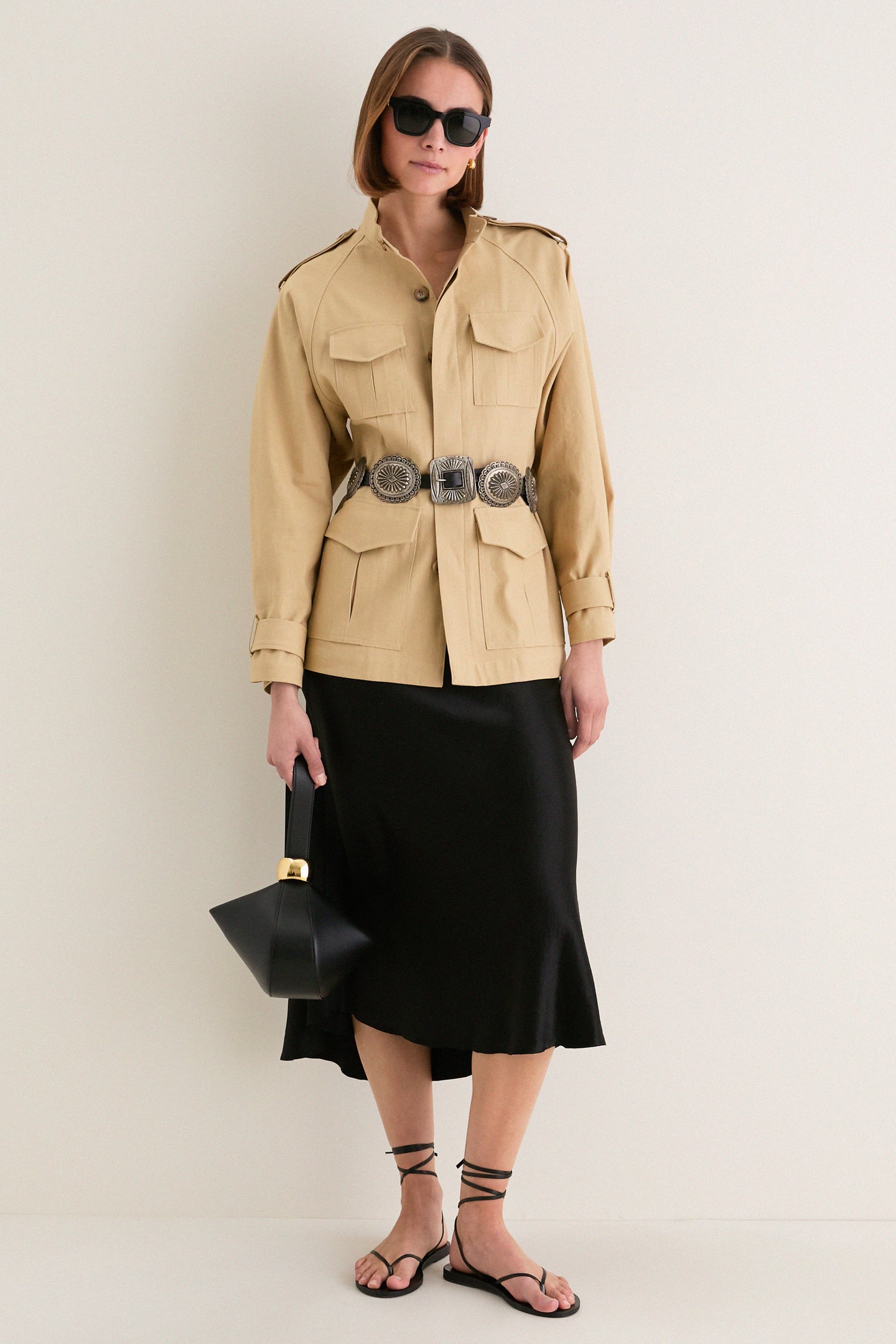 Model wearing the Classic Camel Cotton Field Jacket, cinched with a silver concho belt, paired with a black satin skirt and geometric handbag for an effortless, high-end look.