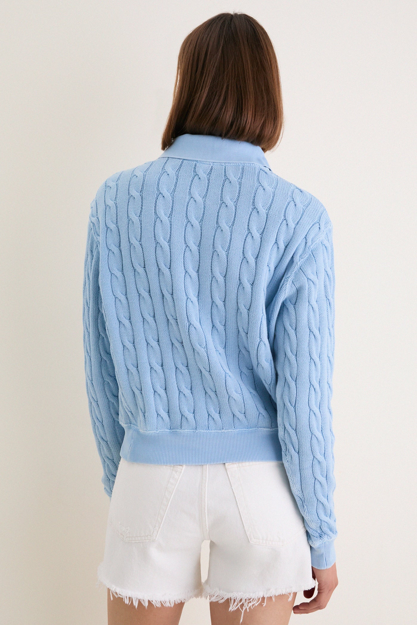 Person wearing Chambray Blue Cotton Knit Polo with classic cable-knit design, polo collar, and ribbed details, paired with high-waisted white denim shorts for an effortless, high-end look.