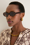 Person wearing Brown Tortoise Lyric Leigh Sunglasses, showcasing their classic oval shape. Effortlessly paired with chunky gold accessories, embodying a fun, high-end style.