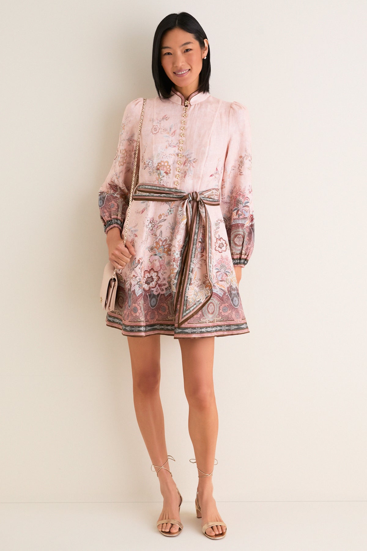 Woman models the classic Pink Paisley Alchemy Button Mini Dress, featuring a floral print, high collar, and sash belt, paired with a beige shoulder bag and sandals.