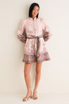 Woman wearing the classic Pink Paisley Alchemy Button Mini Dress with a high collar, floral print, and matching sash, paired with strappy metallic block-heeled sandals.