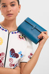 Woman holding a classic Teal Small Soft Clutch by her face, showcasing its fold-over silhouette and handcrafted details, effortlessly blending fun and high-end style.