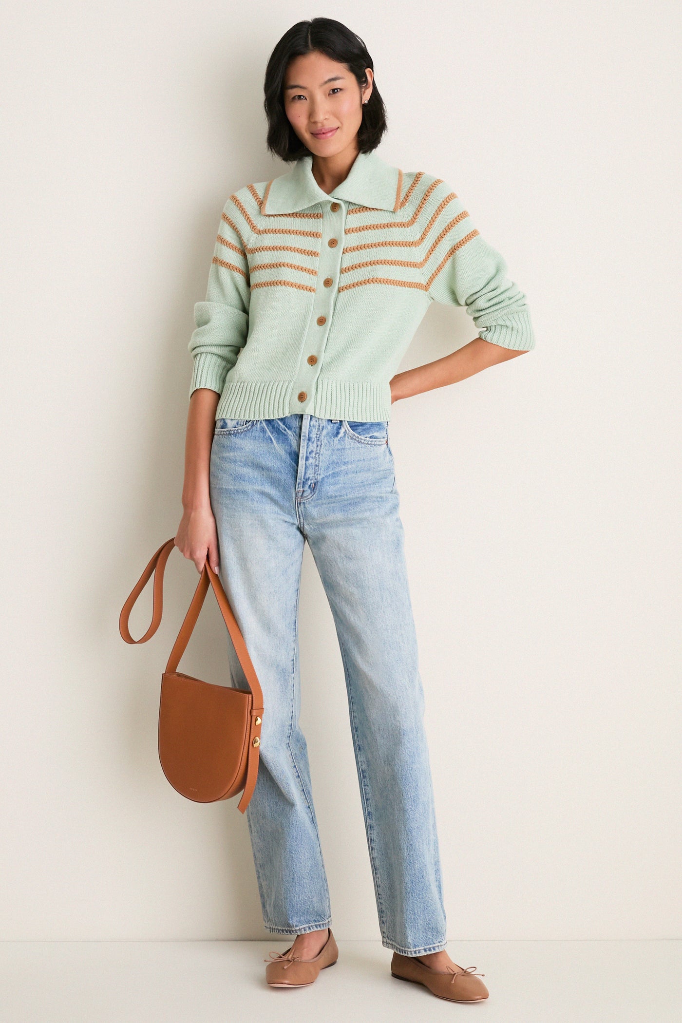 Model in light wash jeans and mint cardigan holds a classic, cognac leather Nigeria crossbody, showcasing its effortless, high-end design with sculptural hardware.