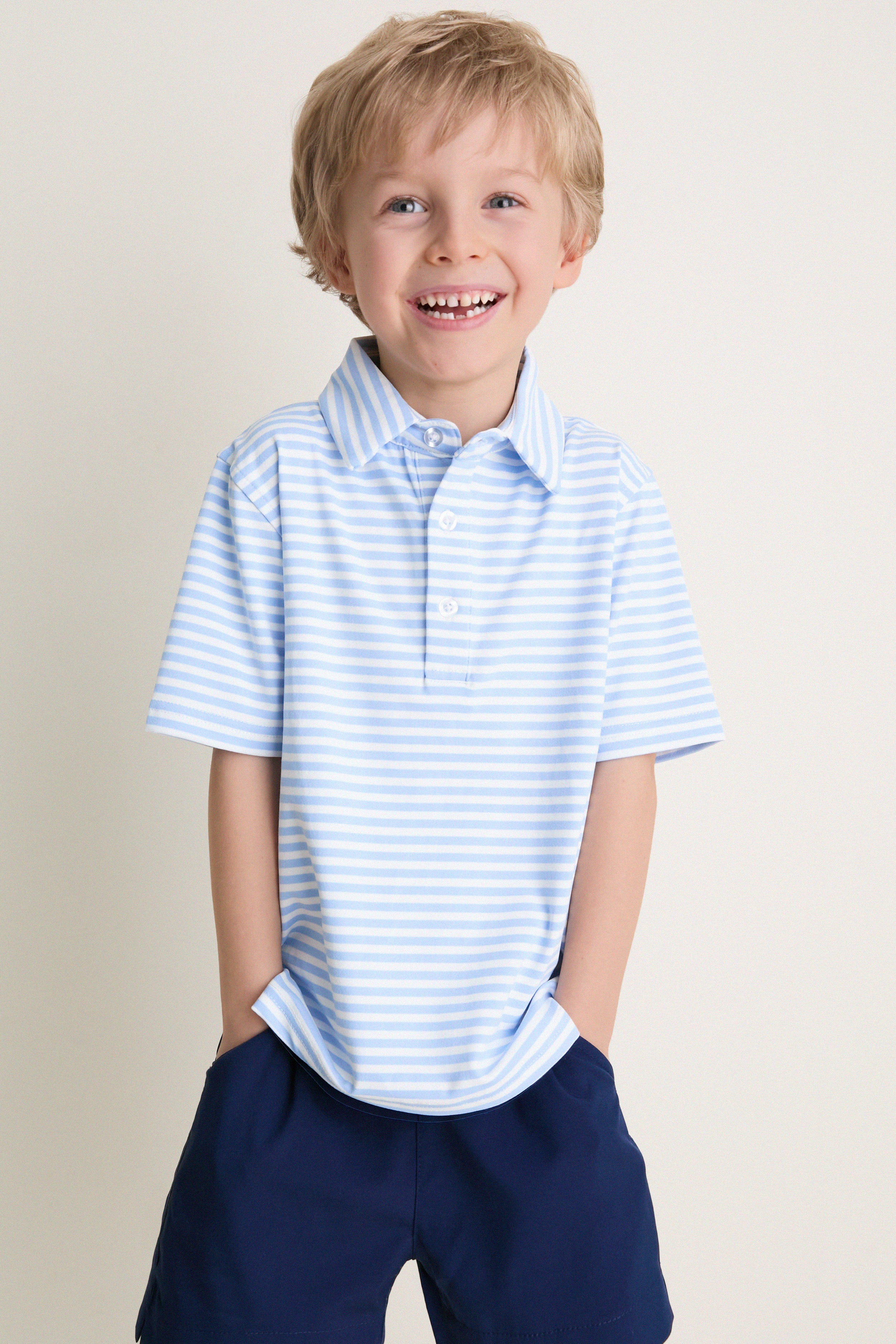 Young boy in classic, effortless Bachelor's Blue and White Le Club Polo, smiling with hands in navy shorts' pockets. Perfect for fun, high-end everyday wear.