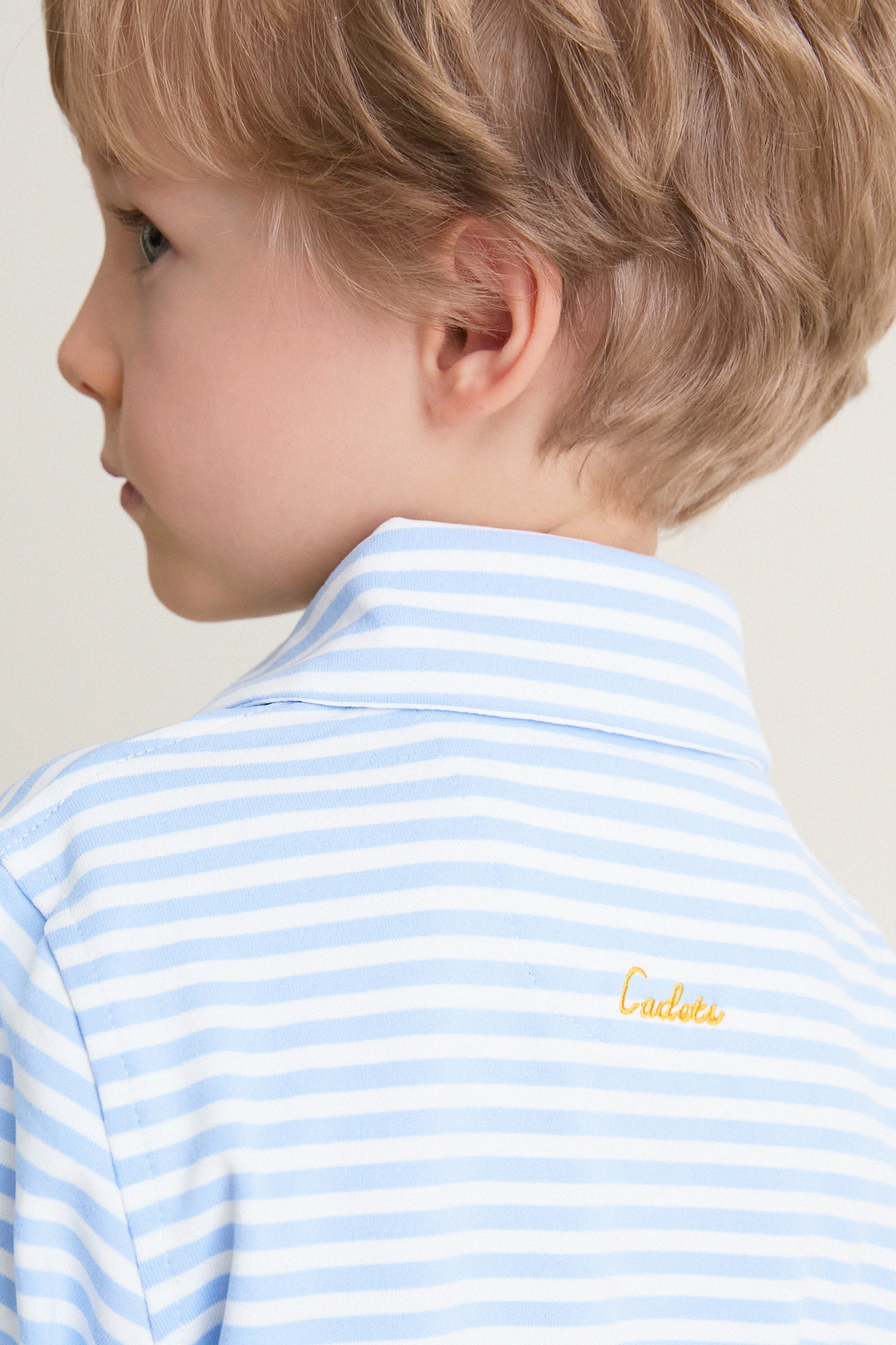Child in a classic, light blue-and-white striped Bachelor's Blue and White Le Club Polo, featuring discreet yellow Cadets embroidery on the back, embodying effortless, high-end style.