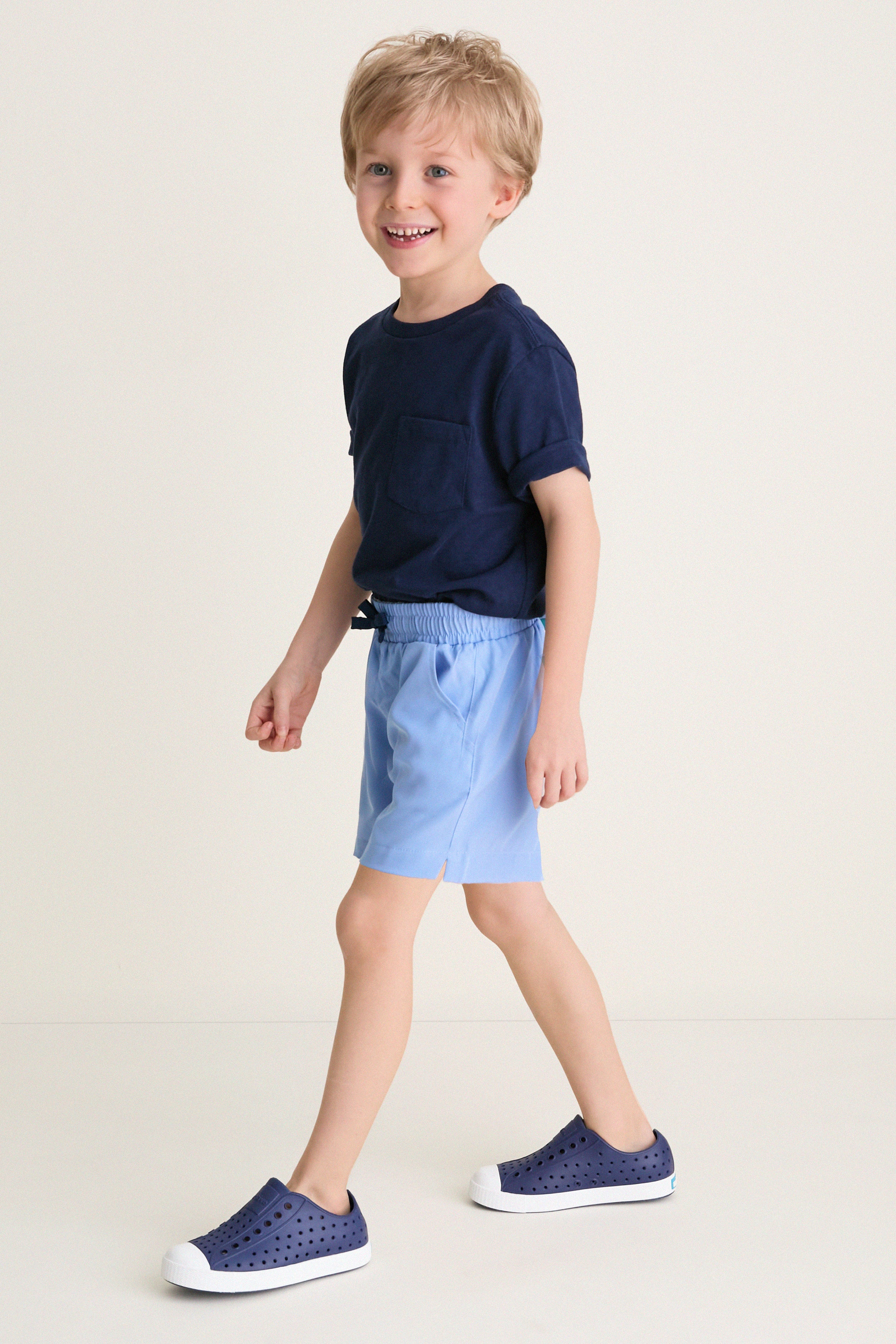 Young boy wearing Sardinia Sky Le Club Short and navy sneakers, mid-step, embodying a classic, effortless style; perfect for high-end, fun everyday wear.