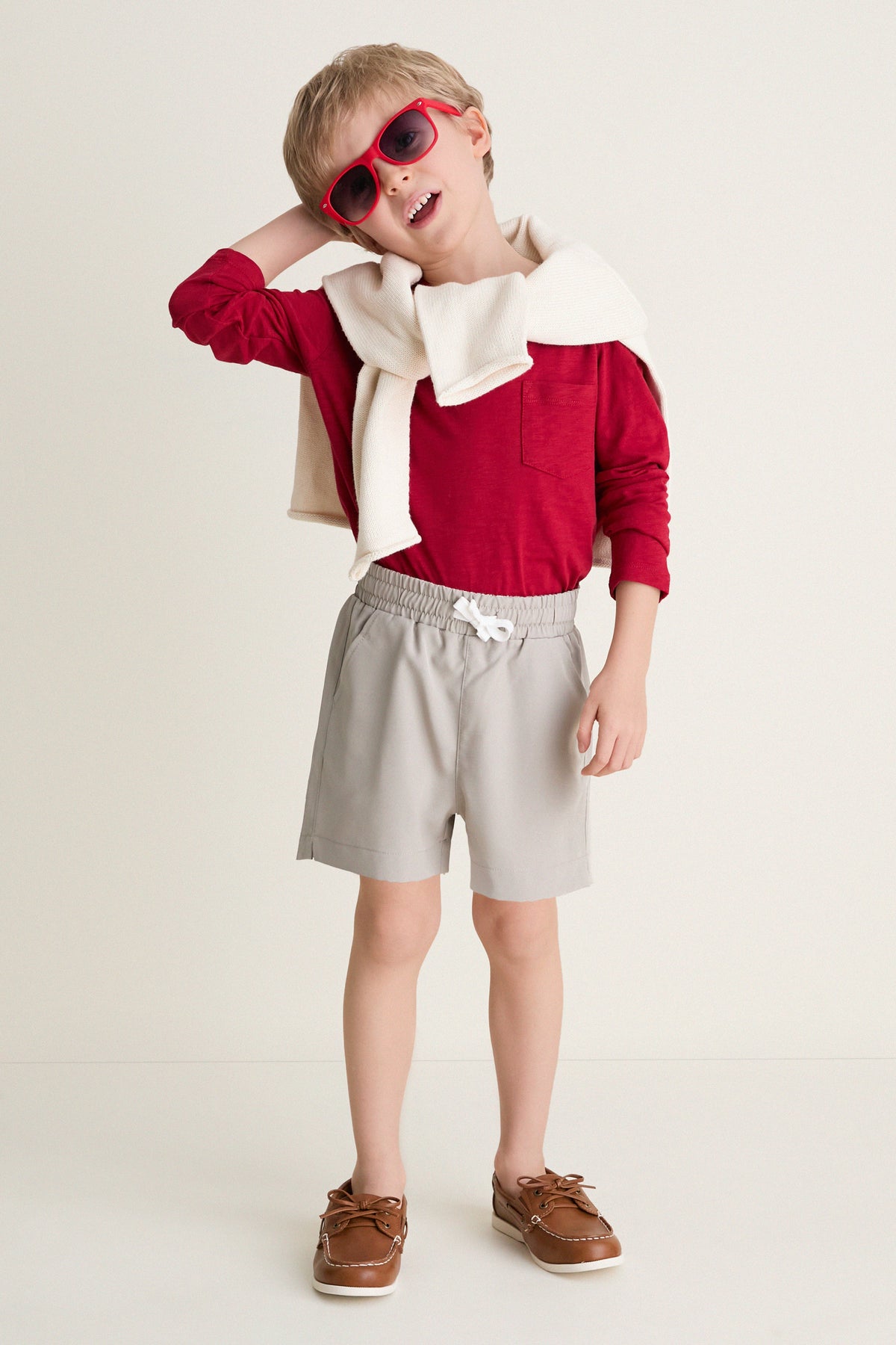 Young boy in Patagonian Putty Le Club Short, styled with a red shirt and cream sweater, embodies classic, effortless style with fun, high-end comfort and versatility.