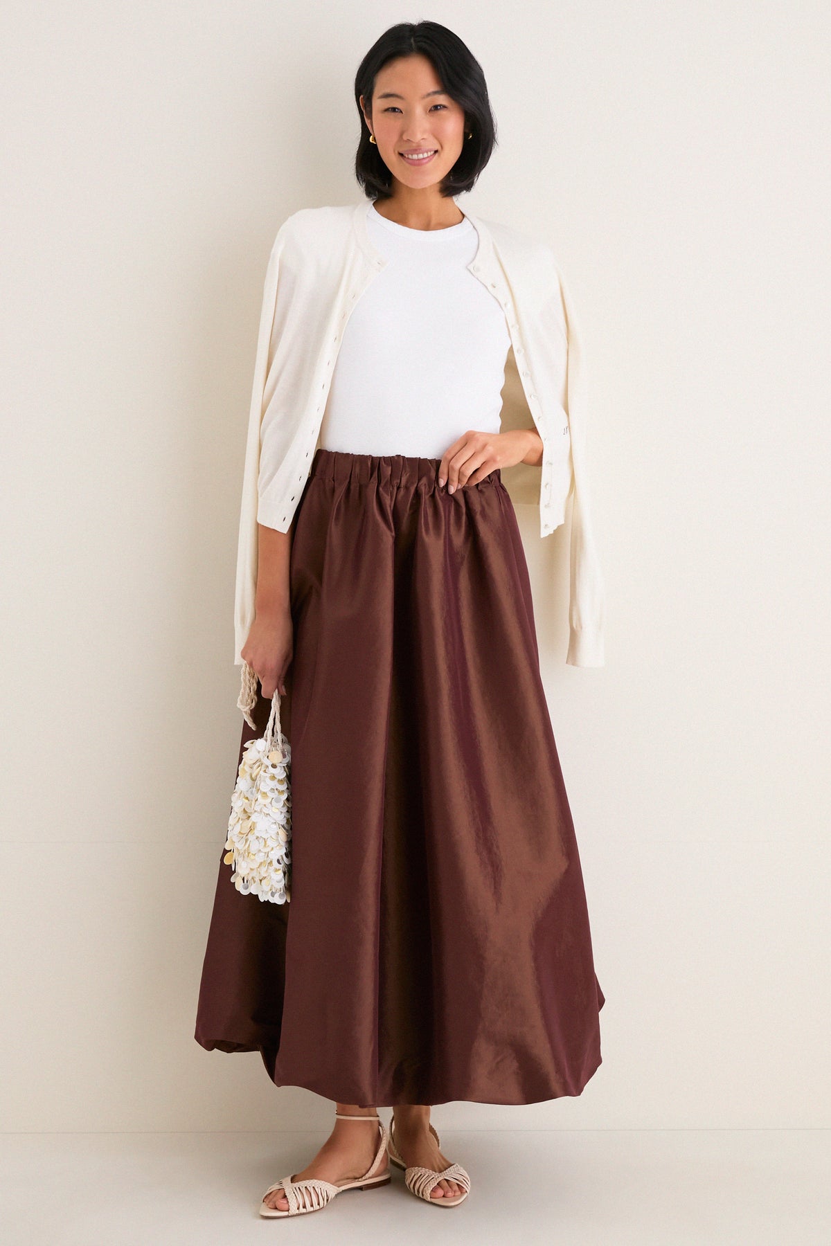Young woman models the classic Chestnut Taffeta Cynthia Bubble Skirt, showcasing its effortless bubble hem and elastic waistband, paired with a sleeveless top and cream cardigan.