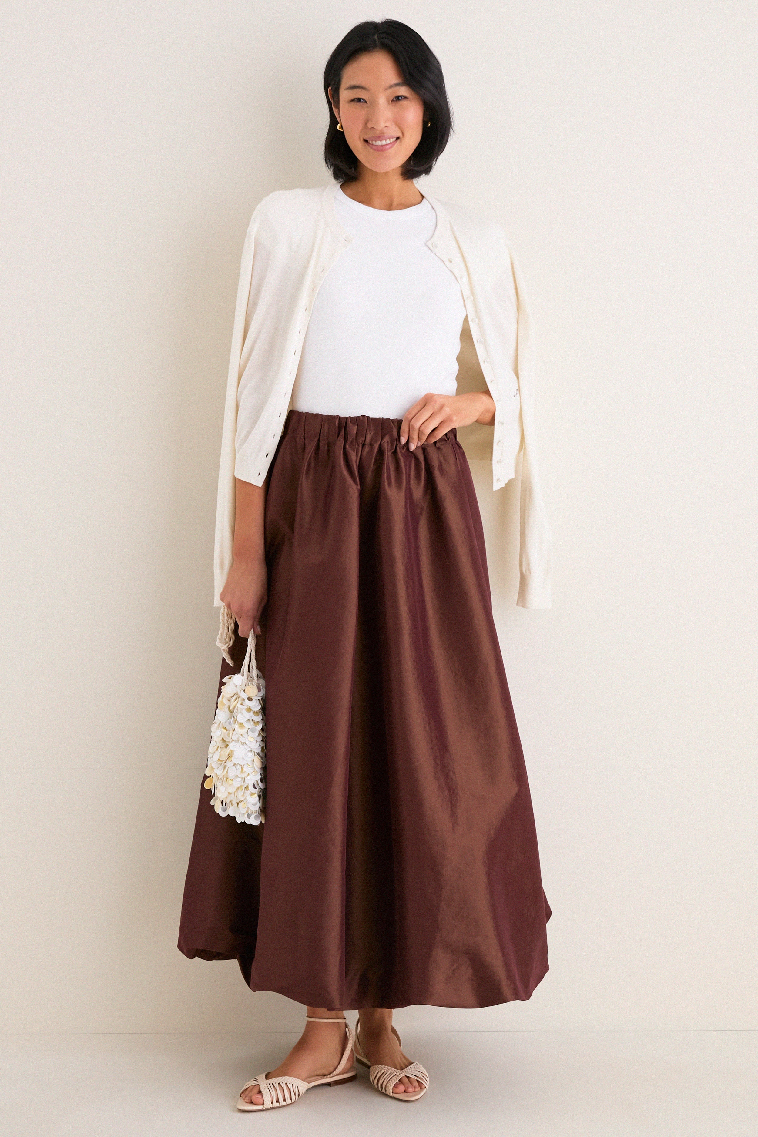 Young woman models the classic Chestnut Taffeta Cynthia Bubble Skirt, showcasing its effortless bubble hem and elastic waistband, paired with a sleeveless top and cream cardigan.