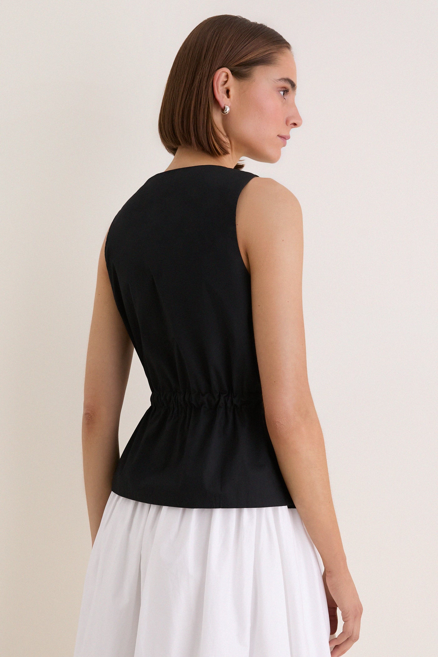 Woman in a sleeveless, classic Black Cotton Poplin Francoise Top with high neckline and cinched waist, exuding effortless sophistication against a simple backdrop.