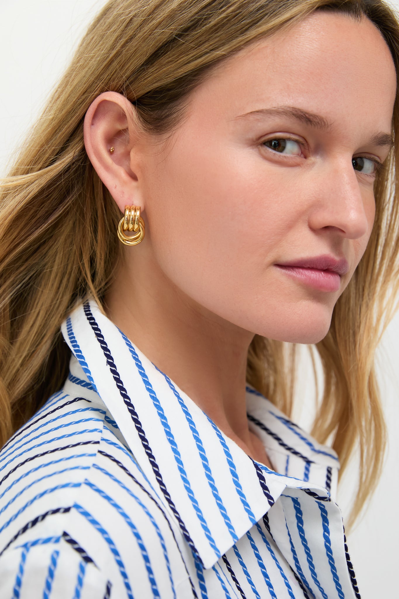 A woman models a blue and white striped shirt and mixed metal earrings by Heaven Mayhem, looking at the camera against a plain background.