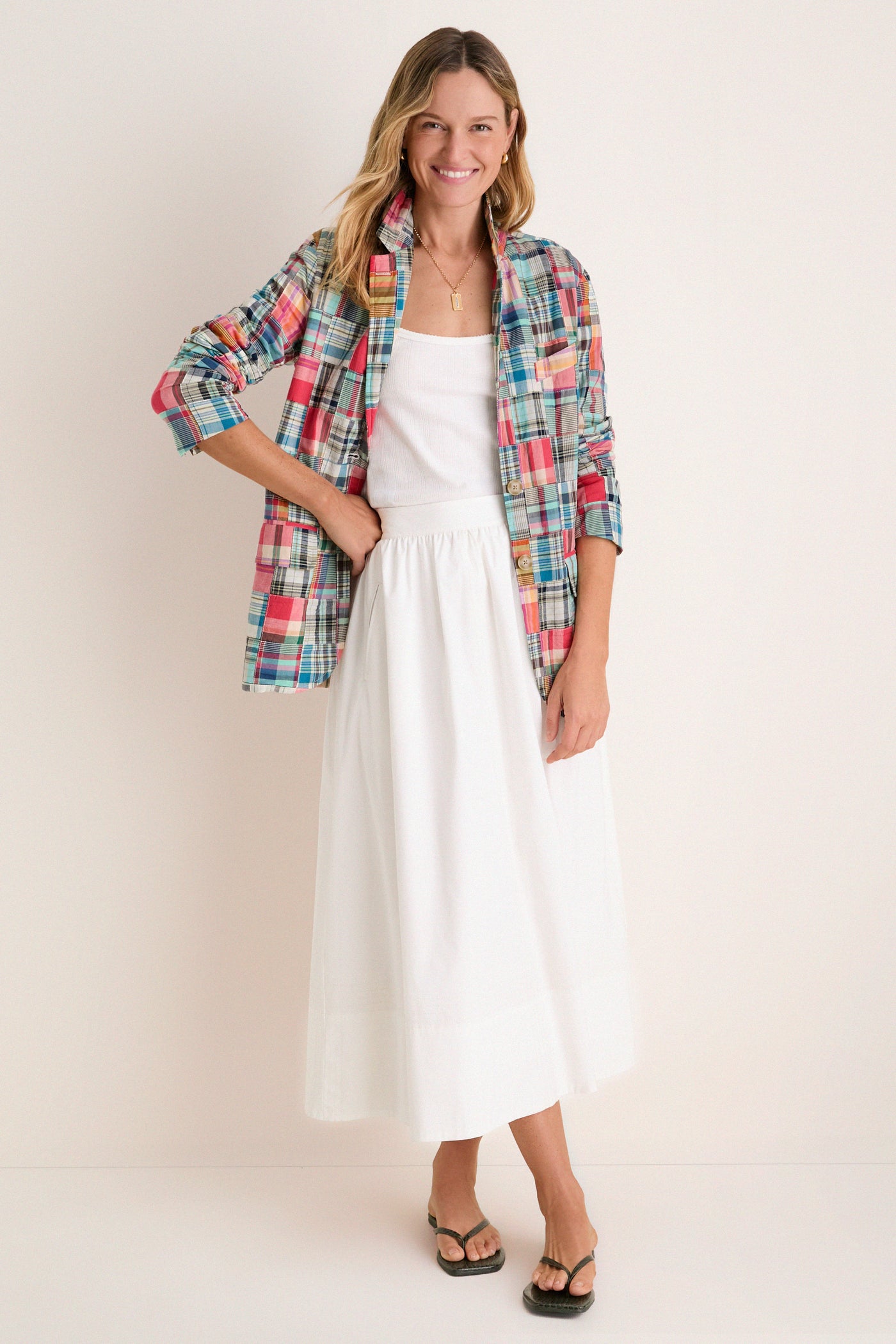 Woman standing in front of a white backdrop in a white tank and skirt and a madras blazer. 