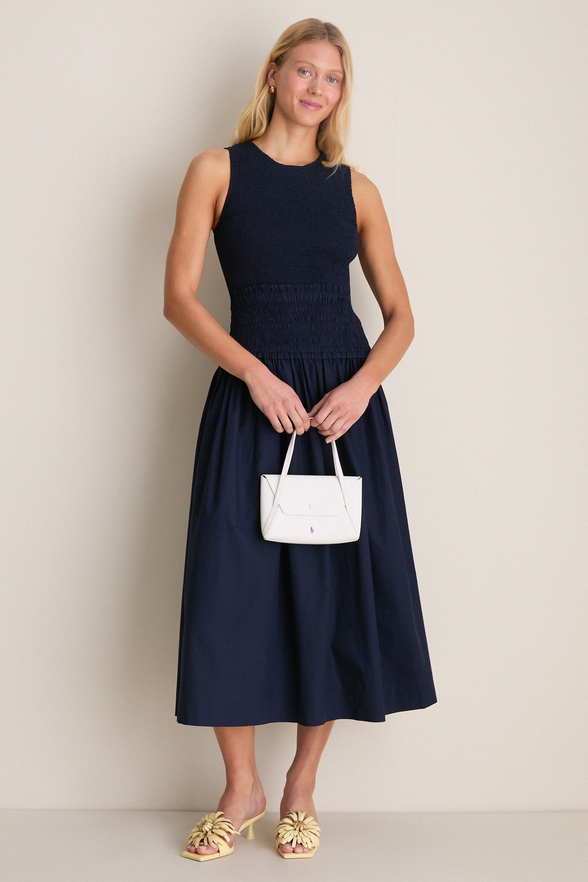 Woman in navy smocked midi dress, holding a white handbag, wears Buttercream Alora Low Heel—classic mule with floral appliqués for an effortless, high-end look.