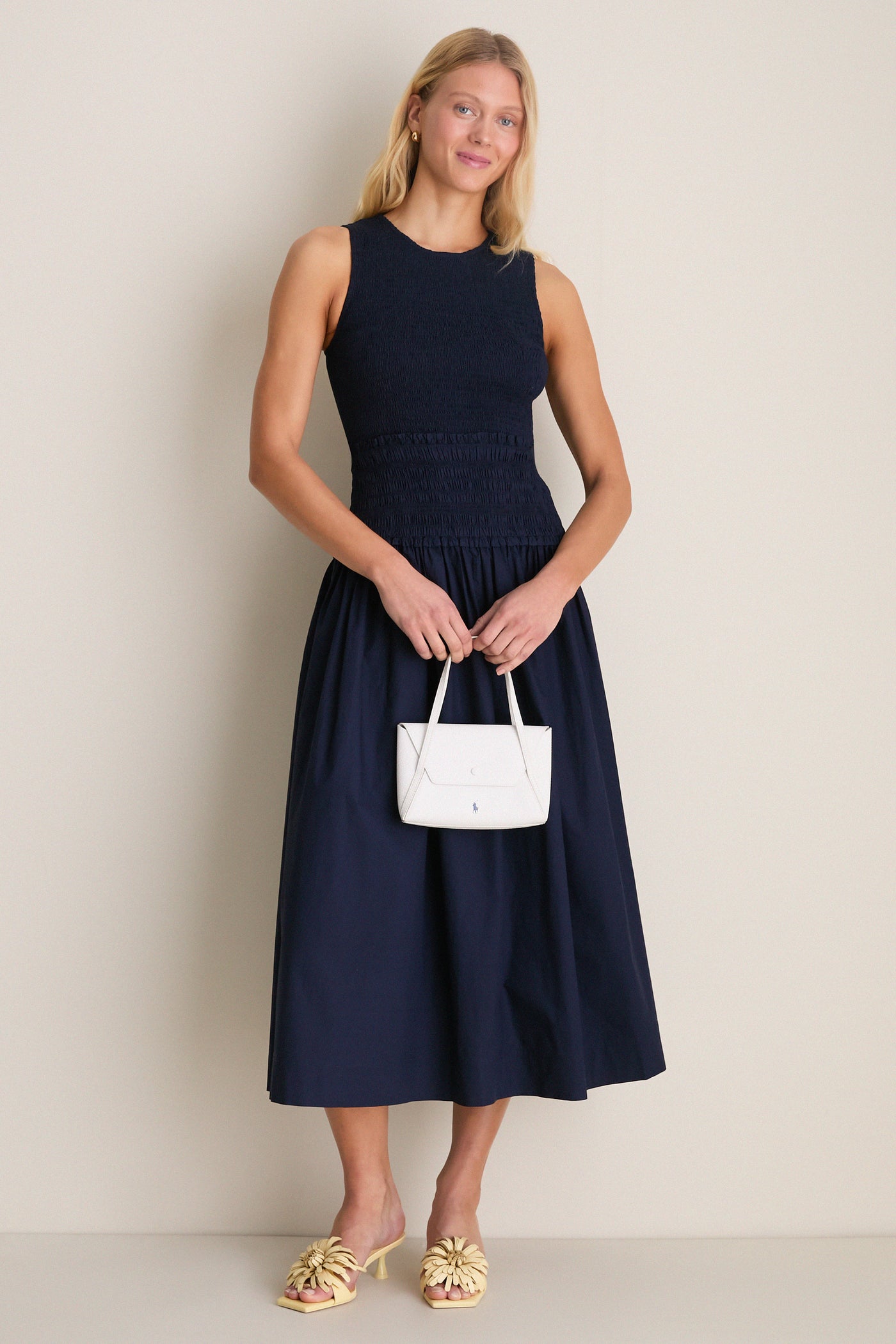 Woman in navy smocked midi dress, holding a white handbag, wears Buttercream Alora Low Heel—classic mule with floral appliqués for an effortless, high-end look.