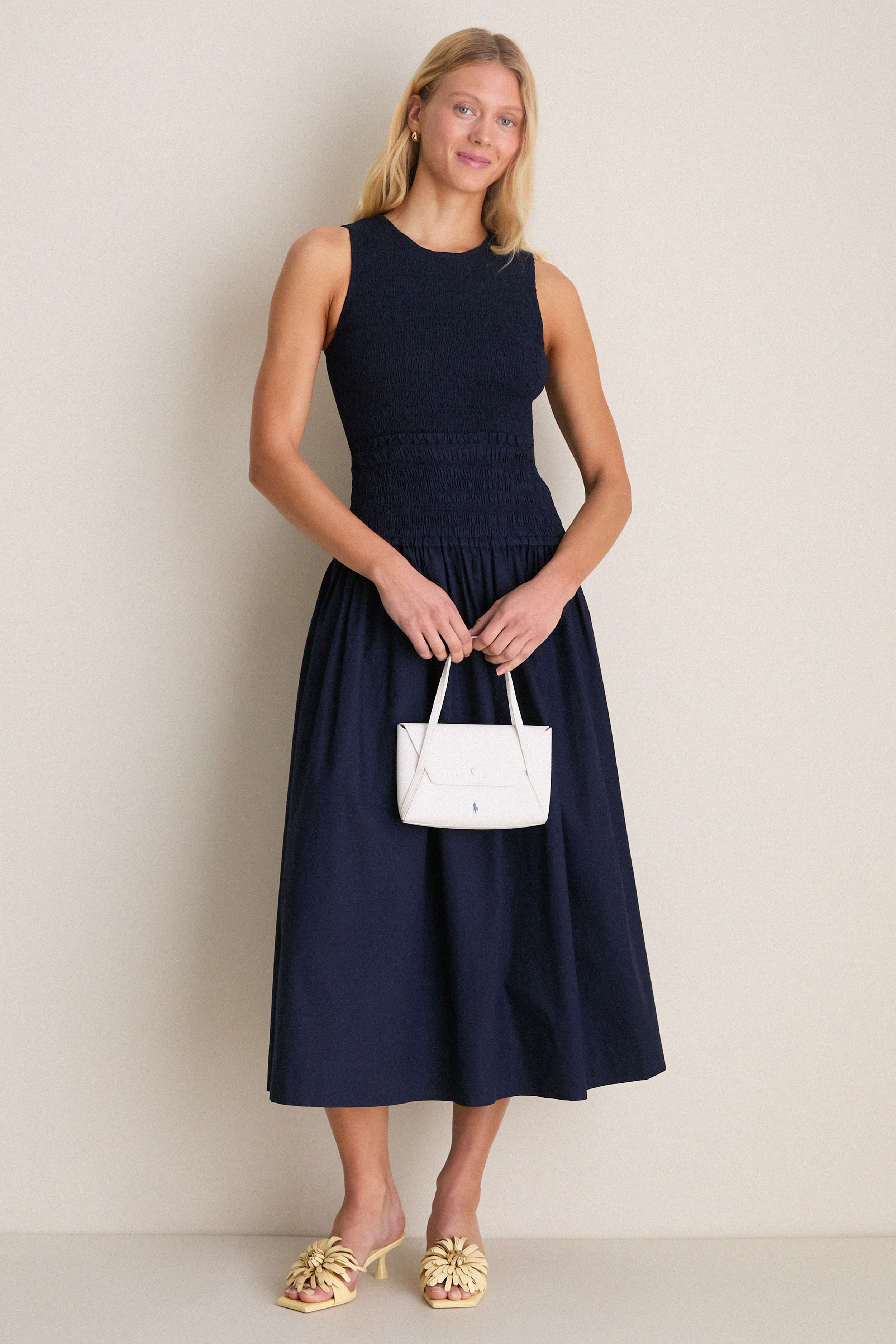 Woman in navy smocked midi dress, holding a white handbag, wears Buttercream Alora Low Heel—classic mule with floral appliqués for an effortless, high-end look.