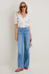 Woman in Coral Jam Jellys, a classic, effortless, and fun look with a cherry-print blouse, light-blue flared jeans, red flats, and a woven straw bag.
