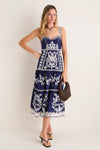 Woman models Blue and White Richilieu Midi Dress with intricate embroidery, sweetheart neckline, tiered skirt; accessorized with wooden-bead handbag and sandals for a classic, effortless look.