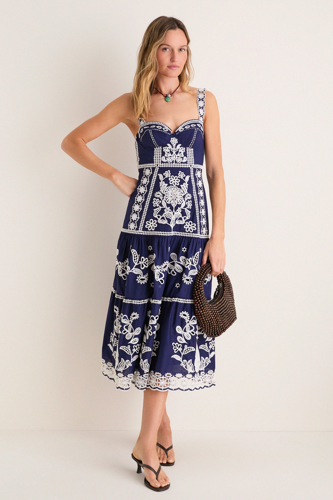 Woman models Blue and White Richilieu Midi Dress with intricate embroidery, sweetheart neckline, tiered skirt; accessorized with wooden-bead handbag and sandals for a classic, effortless look.