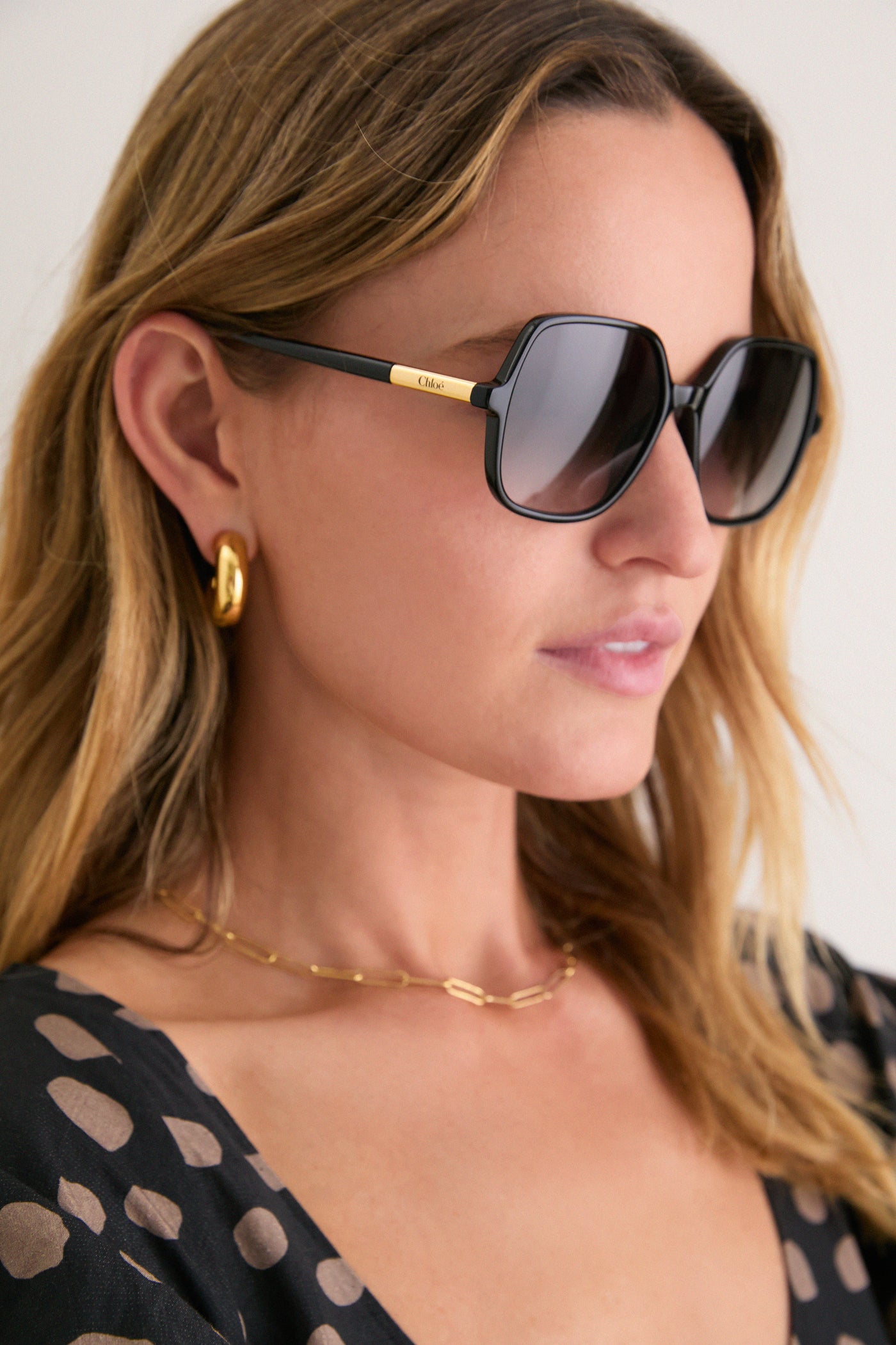 Woman wearing Shiny Black Judy Sunglasses with gold Chloé logo, exuding a classic, effortless style. Features oversized frames paired with chunky gold hoops and chain necklace.