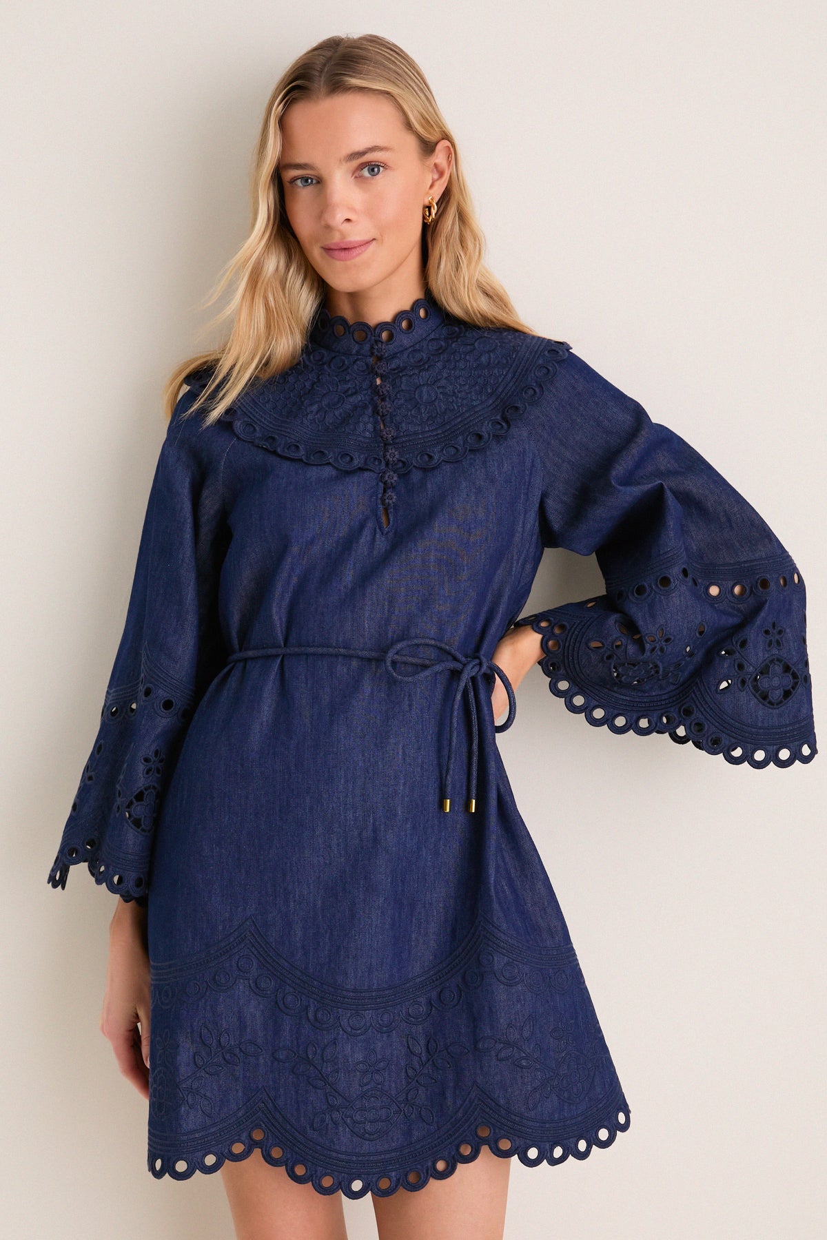 A young woman models the Ink Mahon Broderie Denim Mini Dress, featuring classic scalloped embroidery, effortless bell sleeves, and a fun self-tie belt, embodying high-end, polished style.
