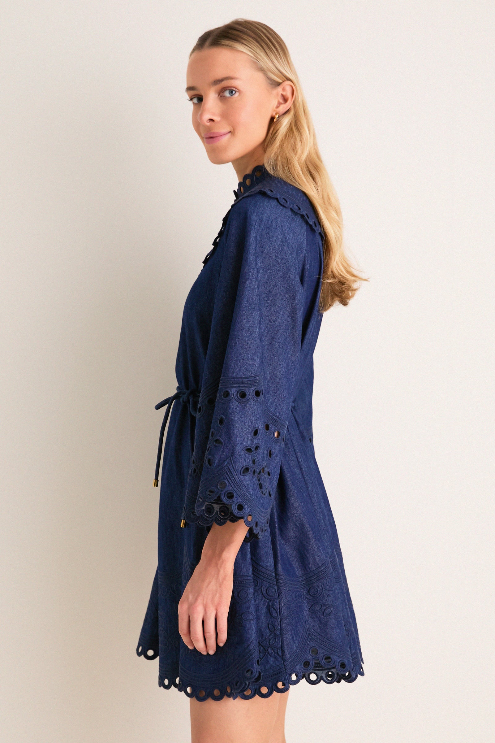 Young woman in profile wearing the Ink Mahon Broderie Denim Mini Dress, featuring scalloped embroidery, eyelet details, and a cinched waist for an effortless, high-end look.