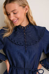 Smiling woman in Ink Mahon Broderie Denim Mini Dress, featuring a classic high scalloped collar, embroidered cuffs, and effortless self-tie waist for a fun, high-end look.