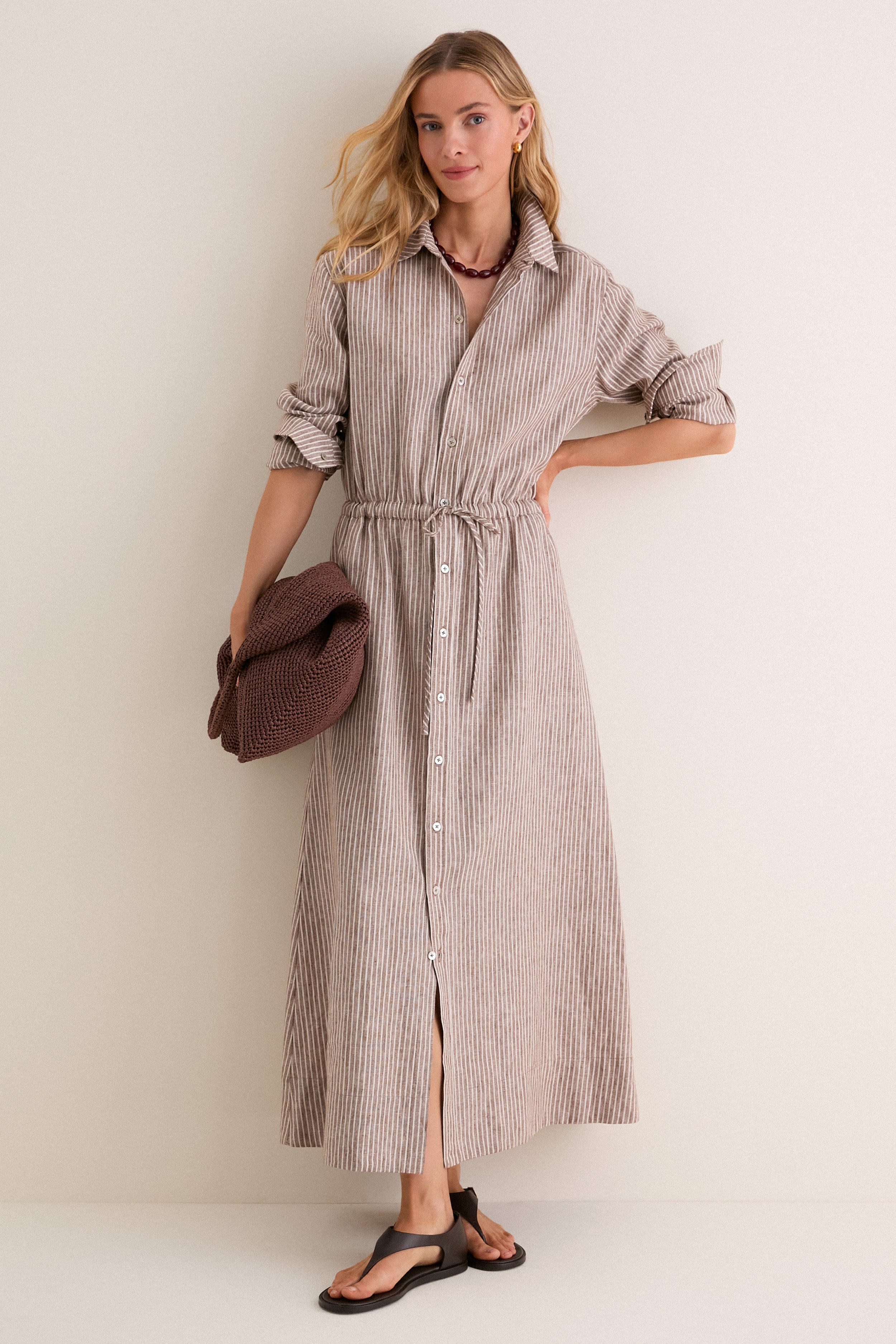 Woman models the classic Cafe Stripe Linen Julien Shirt Dress, featuring an effortless collared neckline, rolled sleeves, and drawstring waist, paired with a chunky-knit sweater and accessories.